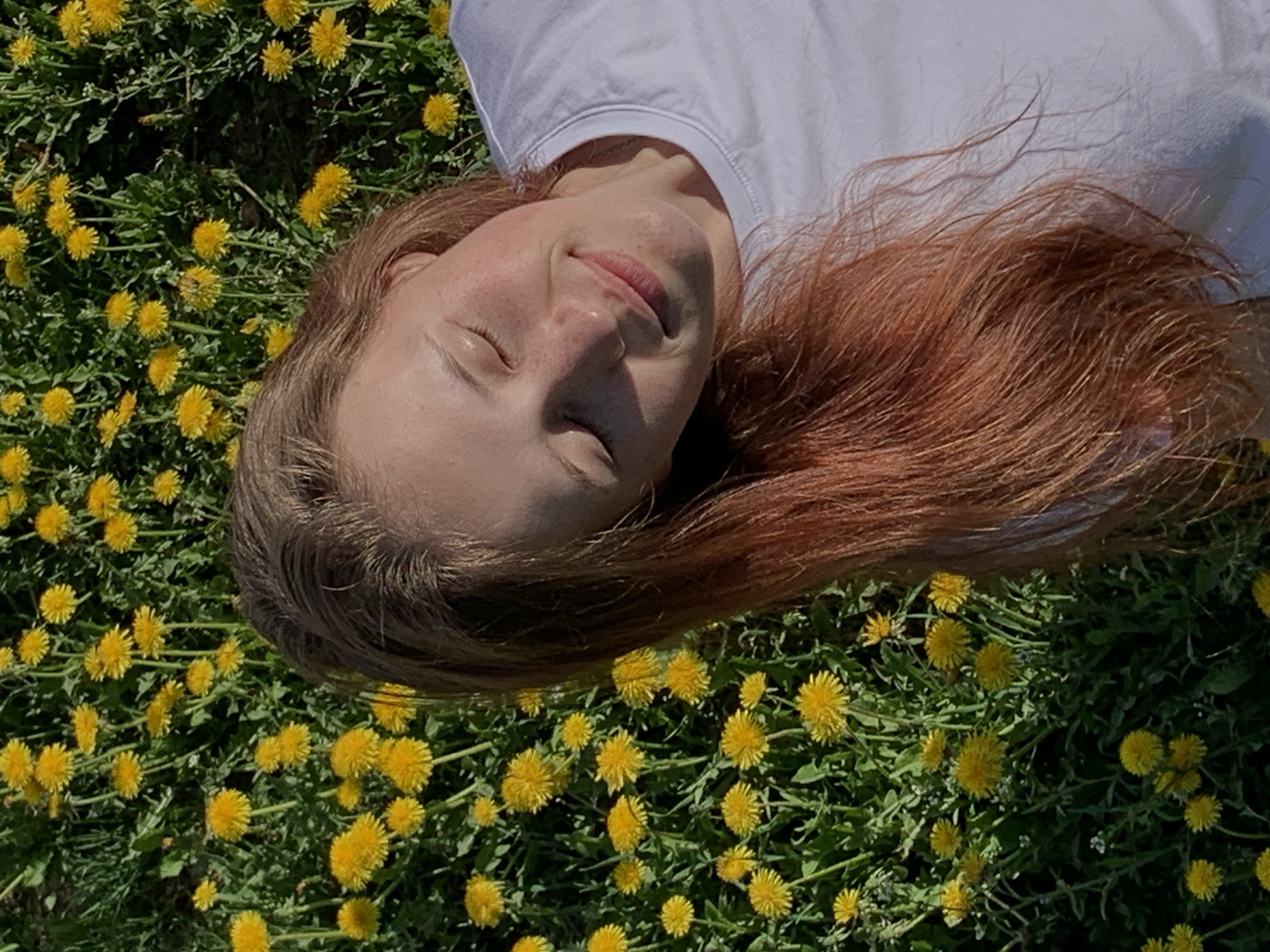 A young woman lies on her back in a field of yellow dandelions. She wears a white shirt. Her hair is reddish-brown and spread around her head on the flowers. Her eyes are closed and she appears to be smiling slightly. The scene is brightly lit, suggesting it is daytime. The exact location within Фоки, Russia, cannot be determined without additional context.