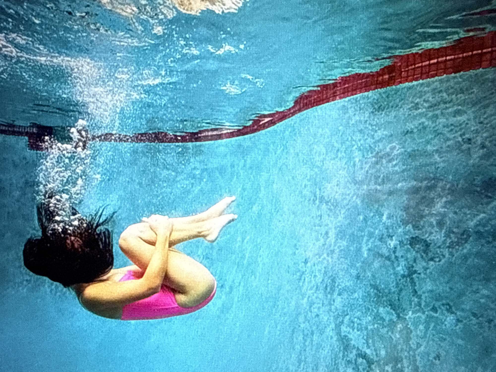 An underwater shot shows a person with dark hair, wearing a pink swimsuit, curled up with their knees to their chest. The person's hair is fanned out around their head, suggesting movement through the water. The background is a bright blue, with shimmering light and ripples. A red lane marker runs vertically down the right side of the frame. The scene is taking place in a swimming pool.
