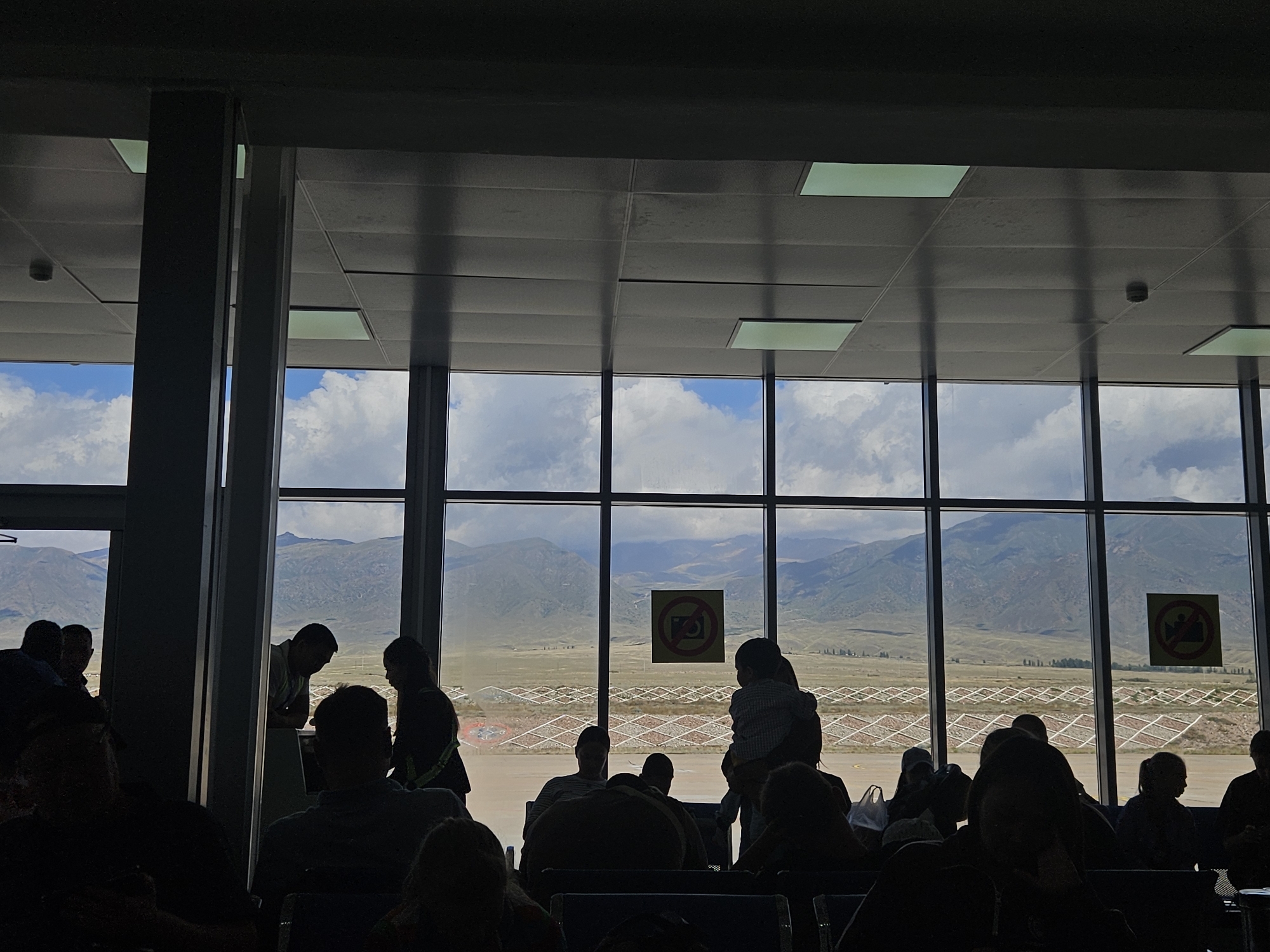 The image depicts an indoor waiting area, likely an airport terminal, with a large panoramic window offering a view of the exterior. Silhouetted figures of people are visible, some seated and others standing, indicating a waiting or transit scenario. Through the window, a vast landscape unfolds, characterized by rolling mountains and a cloudy sky. The outdoor area appears to be an airfield or a similar open space, with geometric patterns visible on the ground, possibly related to runway markings or agricultural fields. Two square signs with a red circle and a diagonal line through an image are present on the window, suggesting a prohibition, likely against photography or videography. The interior is dimly lit, with the exterior view being the primary source of light. The scene is set against the backdrop of the city of Choktal, Kyrgyzstan, known for its mountainous terrain.