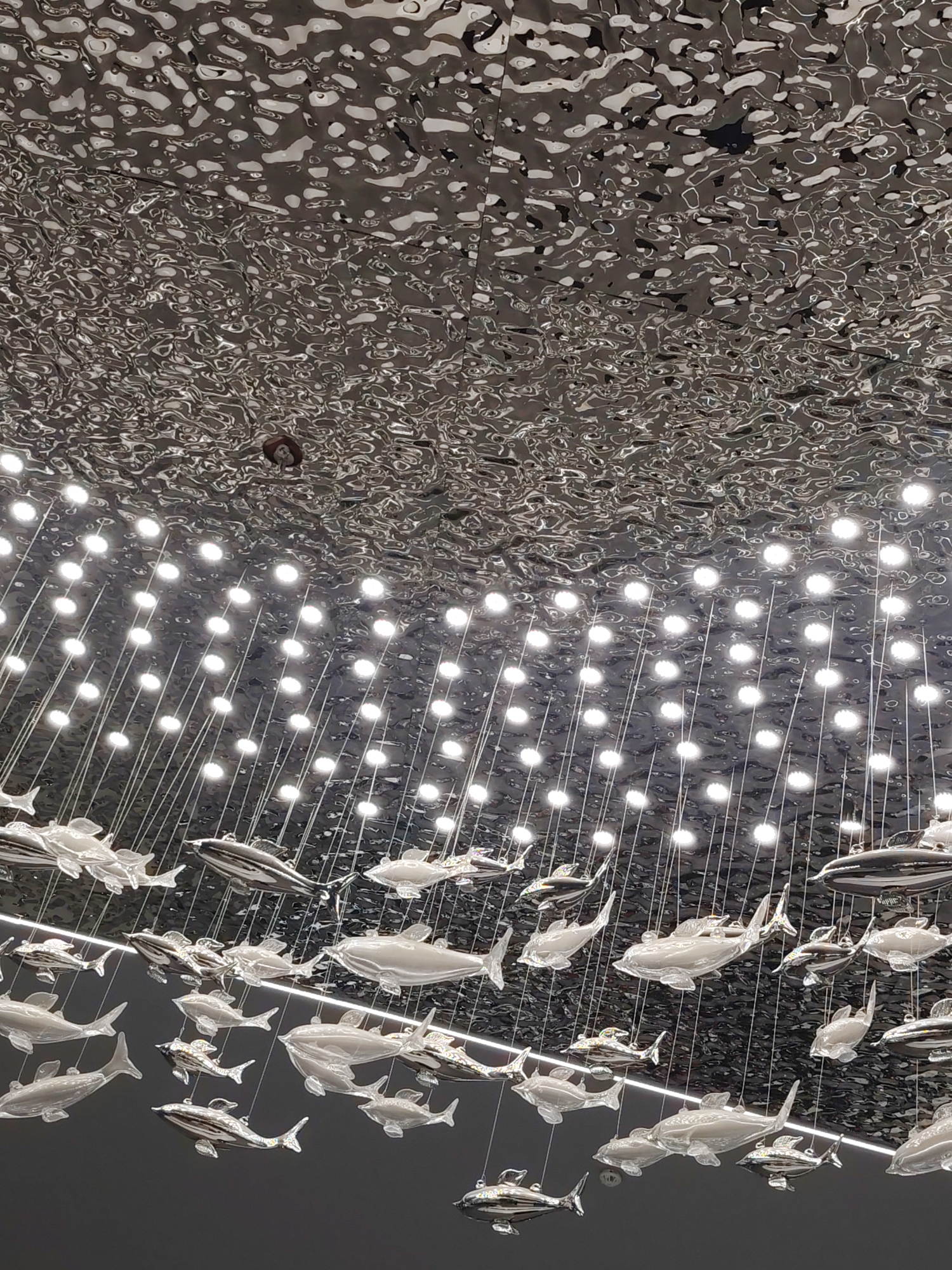 The image shows an indoor space with a highly textured, reflective ceiling that appears to mimic the surface of water. Numerous small, bright lights are suspended from the ceiling in a grid-like pattern, casting illumination downwards. Hanging from thin wires are multiple decorative fish sculptures, varying in size and material, some appearing translucent and others metallic. The overall impression is of an artistic installation, possibly in a modern building or public space. The image does not provide any information about the city of Cholpon-Ata, Kyrgyzstan.