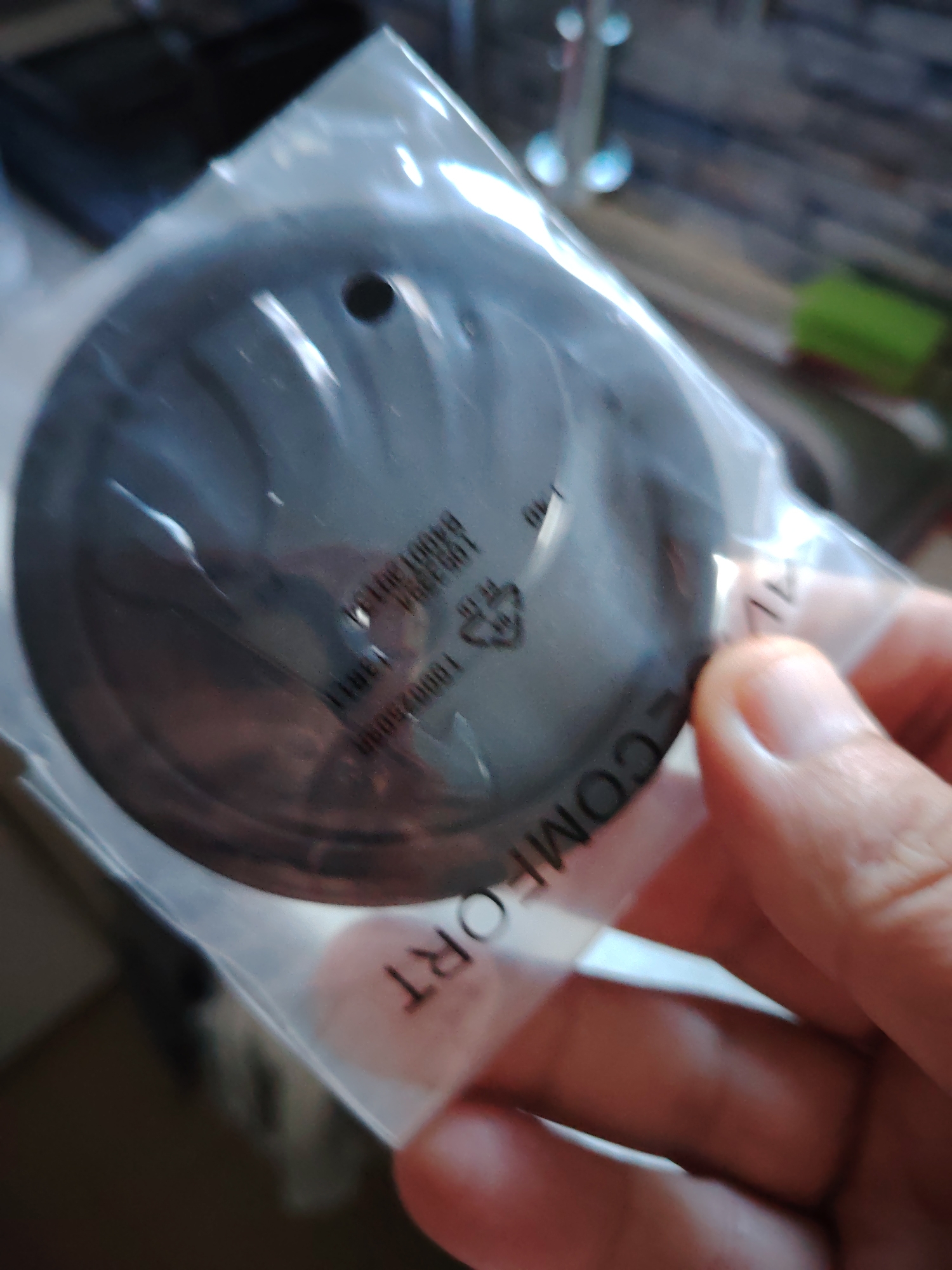 A hand is holding a clear plastic package containing a circular, dark gray object. The object has a hole in the center. Text is visible on the plastic packaging, including what appears to be "COMFORT" and some alphanumeric codes. The background is blurred, showing indistinct shapes and colors that suggest an indoor environment. There are no visible landmarks or cues to indicate a specific location within Huntingdonshire, United Kingdom, or any city.
