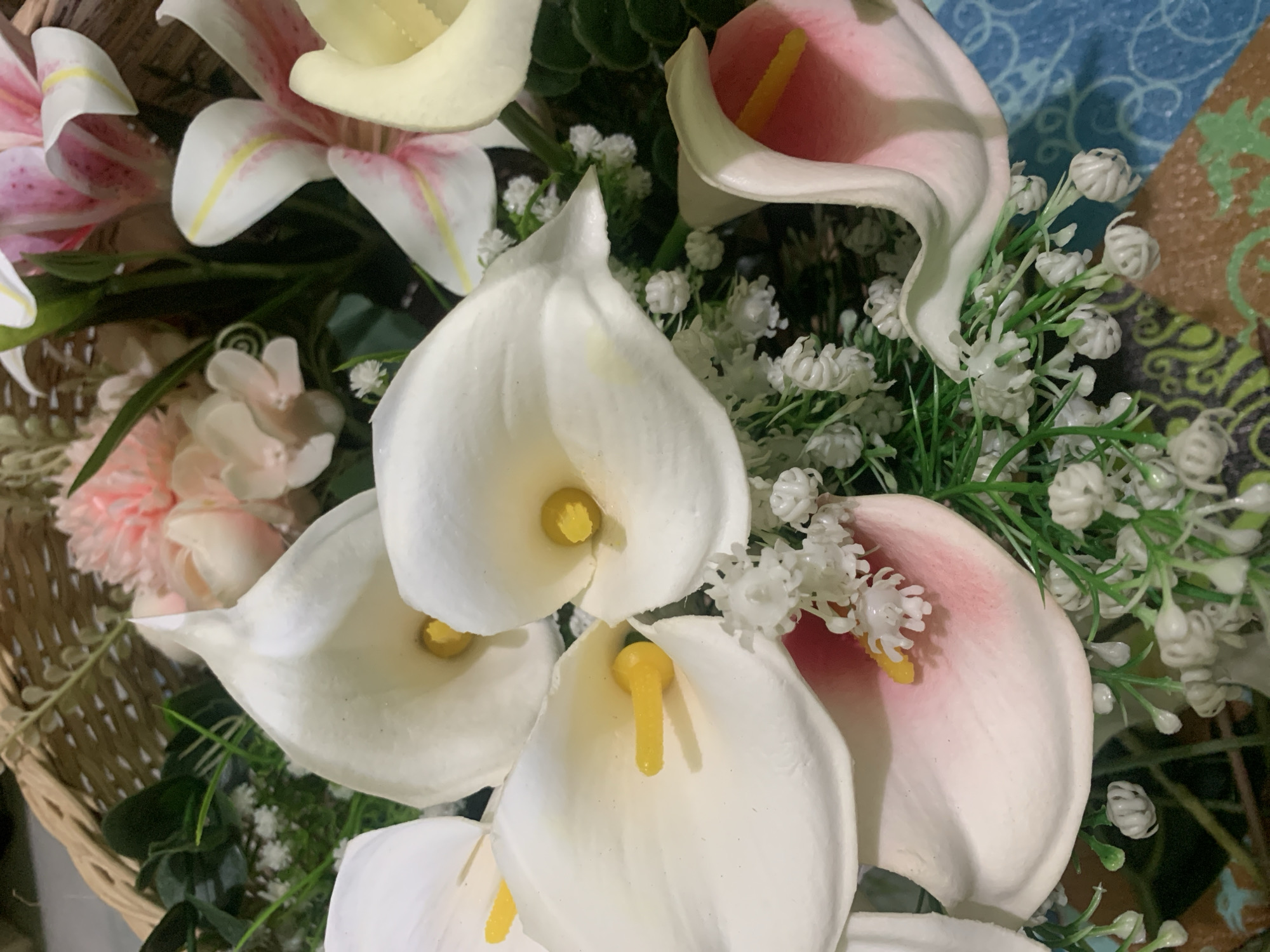 The image shows a close-up of a floral arrangement. The arrangement features several white calla lilies with yellow stamen, some with hints of pink at their edges. Interspersed among the calla lilies are smaller white flowers that resemble baby's breath, and artificial green foliage. In the background, a woven basket can be seen, containing more of the floral elements, including pink carnations and lilies with pink and white petals. The flowers appear to be artificial. The arrangement is placed on a surface with a blue patterned texture. There is no discernible indication of the location being Kuala Pembuang, Indonesia, as there are no landmarks, signage, or environmental cues to suggest this specific geographical setting. The scene is static and appears to be a still life of decorative flowers.