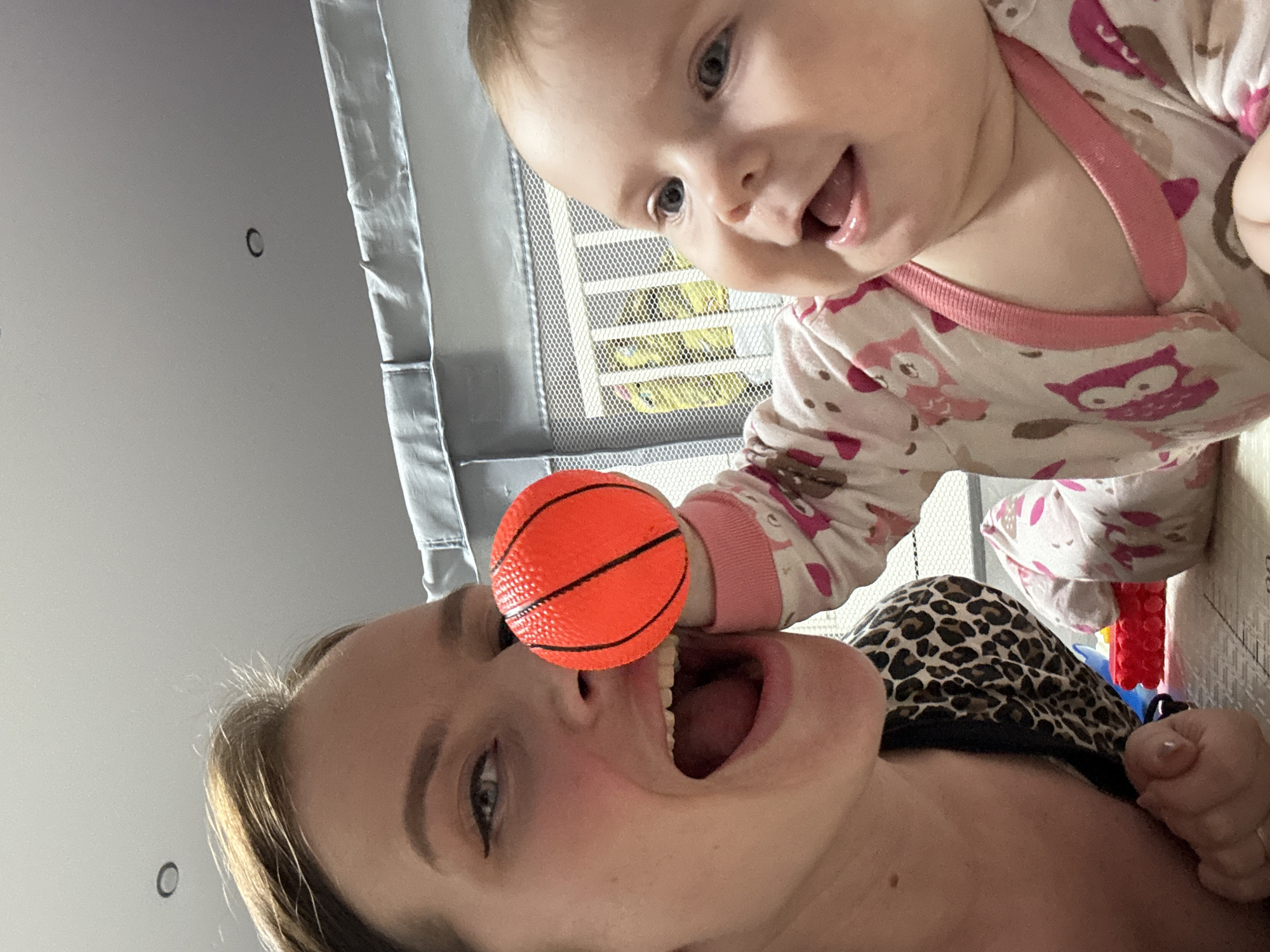 A rotated, close-up indoor shot shows a woman and a baby. The woman is lying down, with an orange miniature basketball positioned in front of her open mouth. She is wearing a patterned top and has light skin and hair. Above her, a baby in pajamas with an owl print is visible, reaching out towards the ball. In the background, there is a grey playpen. There is no information to confirm this scene is in Rusalevka, Russia.