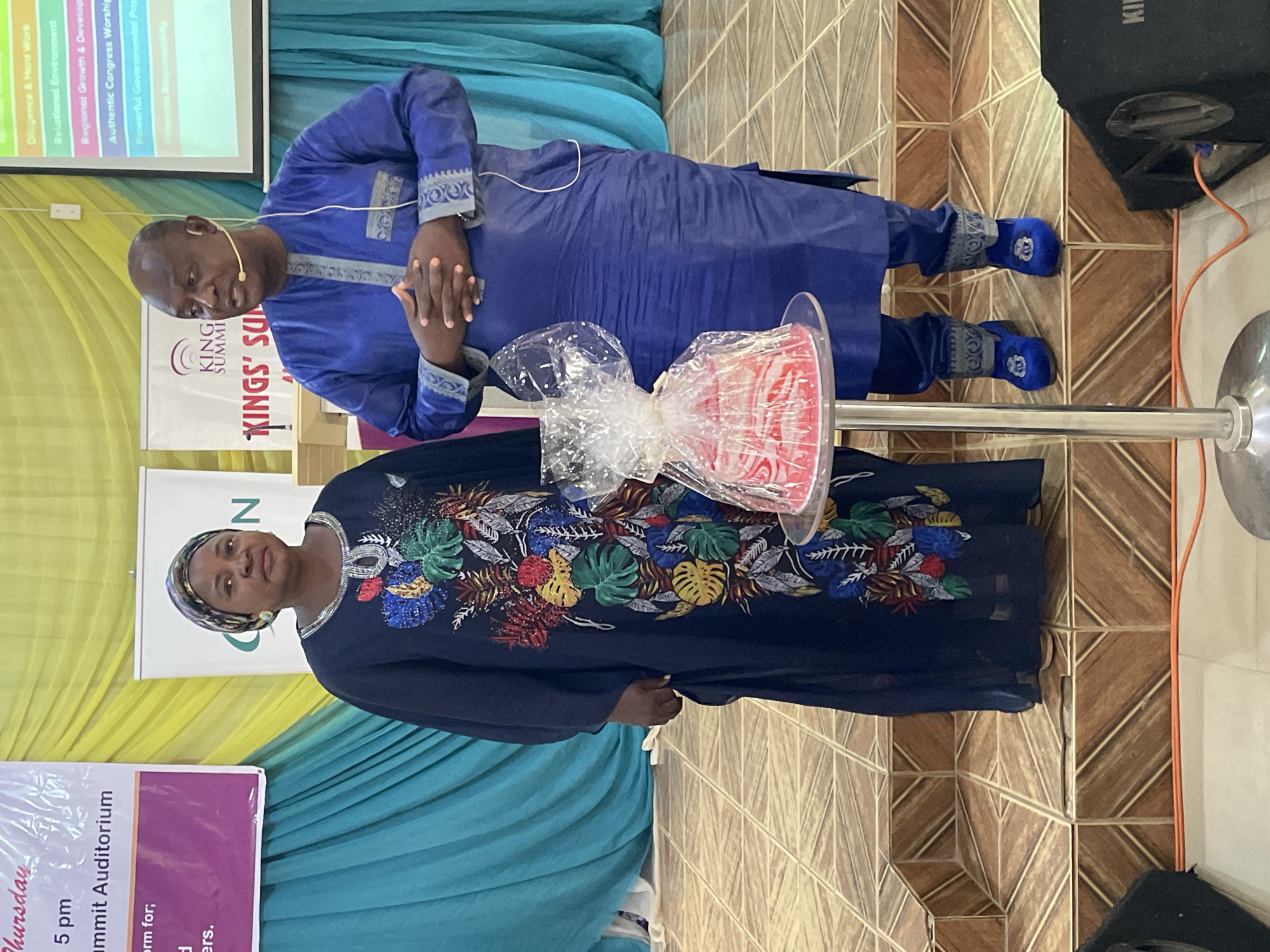The image depicts two individuals, a man and a woman, indoors, likely on a stage or in a presentation setting. The man is wearing a long-sleeved blue garment with ornamental detailing and blue shoes, his hands clasped together in front of him. The woman is wearing a long, dark blue garment adorned with colorful floral patterns, and a headscarf. A gift, wrapped in clear plastic, is positioned between them.
Behind the individuals is a backdrop consisting of teal and yellow curtains. To the left of the frame, there's a partial view of a sign or poster, containing words such as "Auditorium" and information about a Thursday event at 5 pm. A banner is displayed with the text "KING SUMMI" and "KINGS SU". The backdrop includes text mentioning "Diligence & Hard Work", "Authentic Congress Worship", "Regional Growth & Develop", and "Powerful Governmental Pro". The floor has a patterned tile design. The location, Akwanga, Nigeria, is discerned contextually. A black speaker is partially visible on the right.