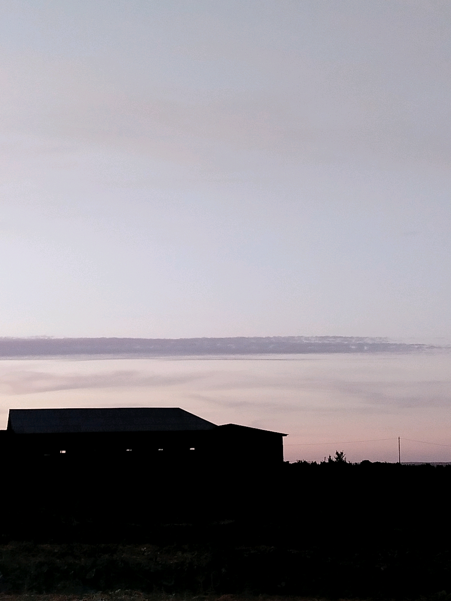 The image depicts a silhouetted building, possibly a barn or warehouse, against a dusky sky. The building is dark and featureless, with small, bright rectangular shapes suggesting windows or openings. A thin, horizontal band of clouds runs across the sky. The foreground is also dark, showing the silhouette of vegetation. The scene appears to be set in a rural area, with the overall mood being calm and subdued due to the low light conditions. The location is stated to be Көлкент, Kazakhstan.
