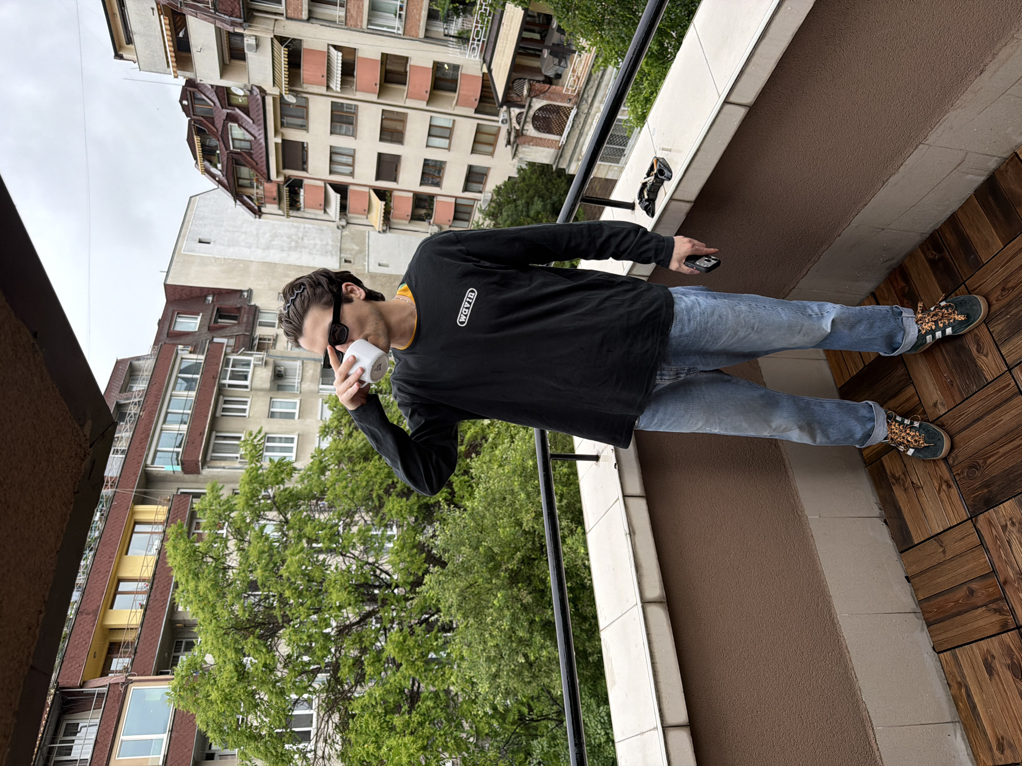 A young man is captured standing on a balcony in a vertical, full-body shot. He is holding a white cup to his mouth with his left hand and a dark-colored smartphone in his right hand. He has dark hair, pulled back by a dark headband or sunglasses, and is wearing sunglasses. His attire consists of a black long-sleeved t-shirt with a yellow accent at the collar and a white oval logo on the chest, which reads "WAVYN". He is also wearing light blue jeans and black sneakers with distinctive leopard-print laces.
The balcony features a patterned brown tiled floor, resembling wood planks, on the right side, and a lighter, textured brown surface closer to the white balustrade on the left. A black metal railing defines the edge of the balcony.
The background reveals a dense urban setting with several multi-story apartment buildings under an overcast sky. The buildings display varied architectural styles, colors, and window configurations, some featuring balconies and external blinds. Vibrant green trees are visible, interspersed among the structures and below the balcony level, suggesting an inner-city green space. The scene depicts a daytime moment in Sofia, Bulgaria, with the man appearing to be casually observing his surroundings or taking a break.