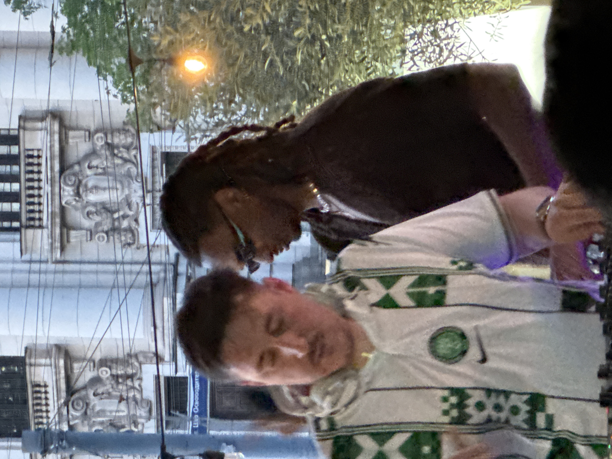 The image depicts two men standing together outside. The man in the foreground is wearing a white hooded sweatshirt with a green Celtic football club logo and green geometric patterns. The man behind him is wearing a black shirt, sunglasses, and has long braided hair. The location appears to be in Sofia, Bulgaria. A building with ornate details and a street sign with Cyrillic text ("Цар Освободител") is visible in the background. A street light is visible above.