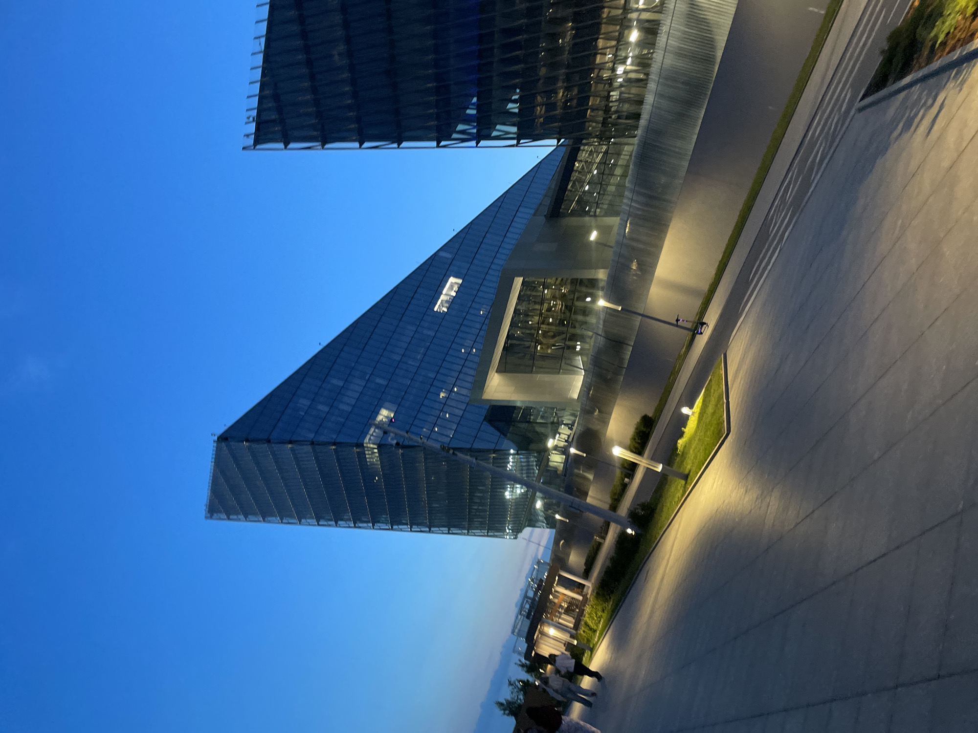The image shows the exterior of the Gazprom Tower in Saint Petersburg, Russia, at dusk. The building, characterized by its striking triangular shape and glass facade, is illuminated by interior lights and exterior streetlights. A pathway, paved with grey tiles, leads away from the viewer, with a well-maintained strip of grass and modern lampposts along its edge. In the background, several people are walking on the pathway. The sky is a deep blue, transitioning to lighter hues near the horizon, indicating twilight. Another glass building, with a darker facade, is visible to the right of the Gazprom Tower. The overall scene depicts a modern urban environment at the end of the day.