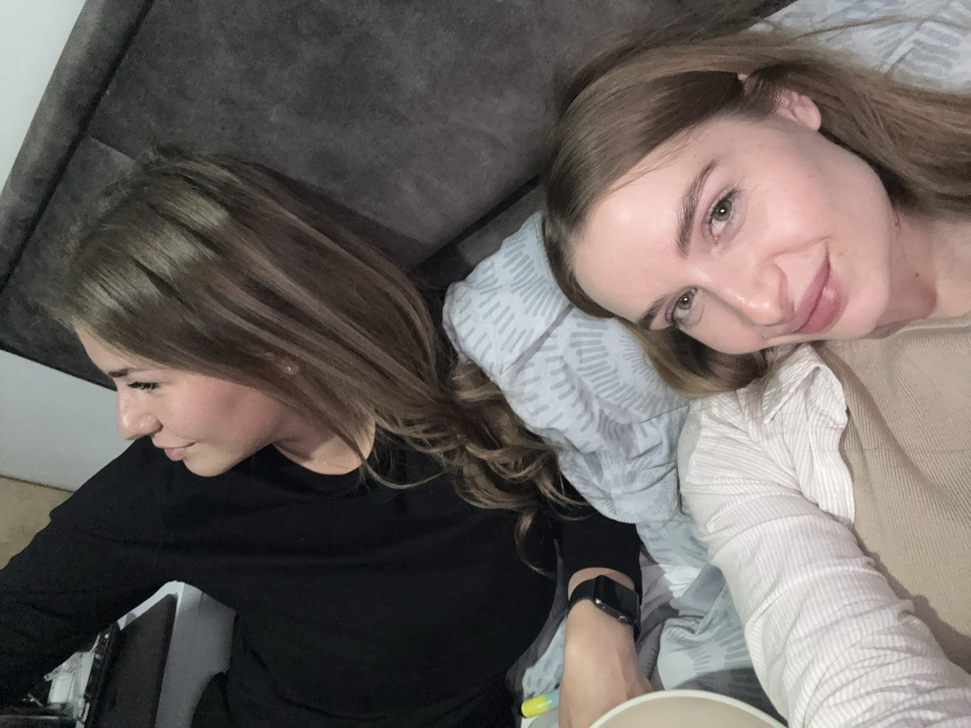 Two adult females are depicted reclining indoors, likely on a bed or sofa. The female in the foreground, positioned bottom-right, has light brown hair, fair skin, and is looking directly at the camera with a slight smile. She wears a beige top layered over a white striped shirt. The second female, positioned top-left, is seen in profile, looking upwards and to the left. She also has light brown hair, fair skin, and wears a black long-sleeved top, with a dark smart-watch visible on her left wrist. They are resting on light-colored bedding, with a patterned light blue and grey pillow beneath the second female's head. A dark grey upholstered backrest or headboard is visible behind them. A yellow and light blue cylindrical object is near the second female's left arm, and a light-colored cup or bowl is partially visible in the bottom-left corner. The indoor setting lacks specific features to identify a precise location within Budva, Montenegro.