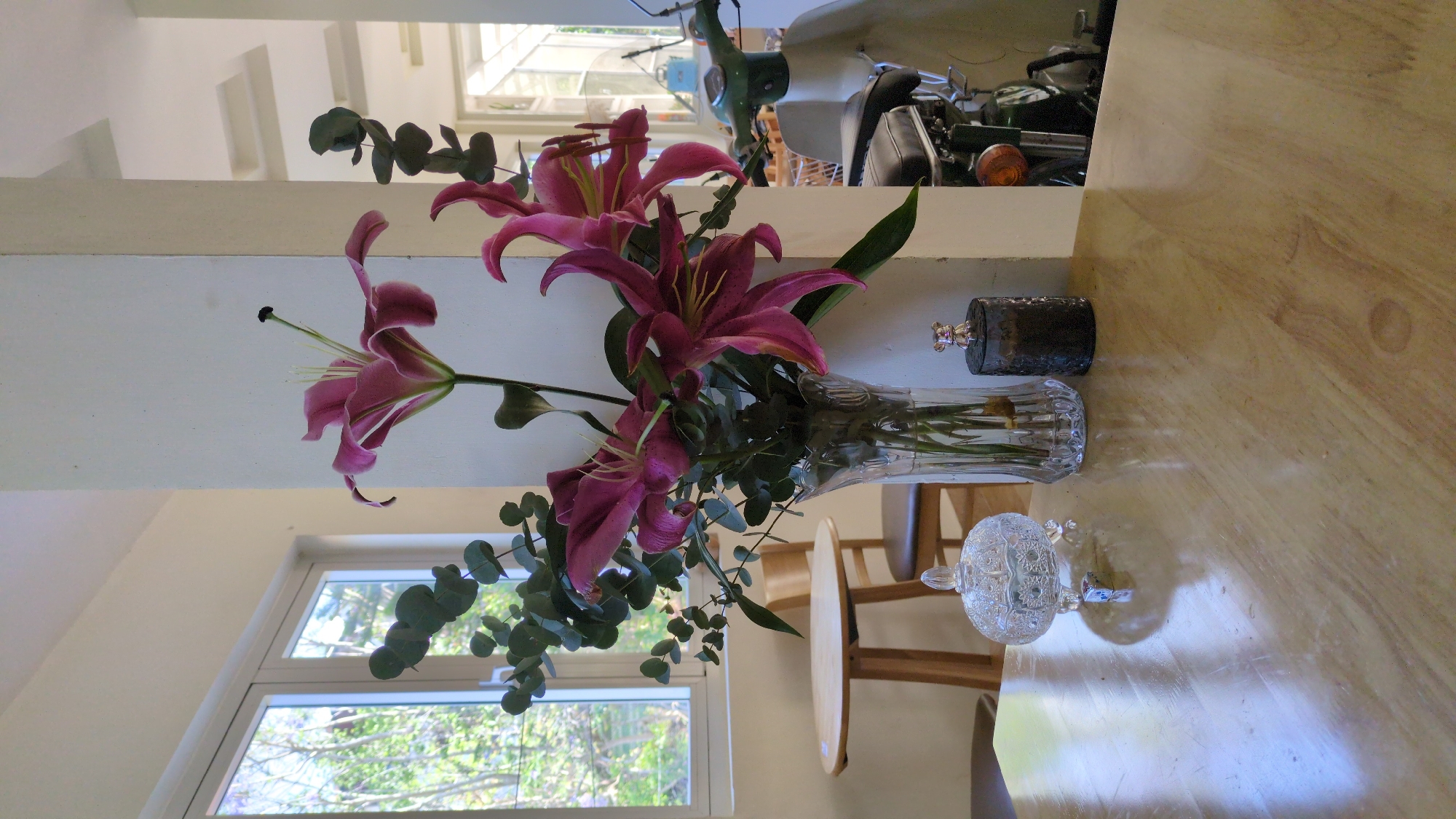 On a light brown wooden surface, a clear vase holds purple lilies and eucalyptus branches. Next to the vase is a small, dark, cylindrical container with a decorative top, and a crystal container with a lid. Behind the vase, a light-colored countertop is visible. Further back, a motorcycle and green scooter are partially in view. A window showing greenery outside is to the left of the vase. The location is Cam Ly, Đà Lạt, Vietnam.