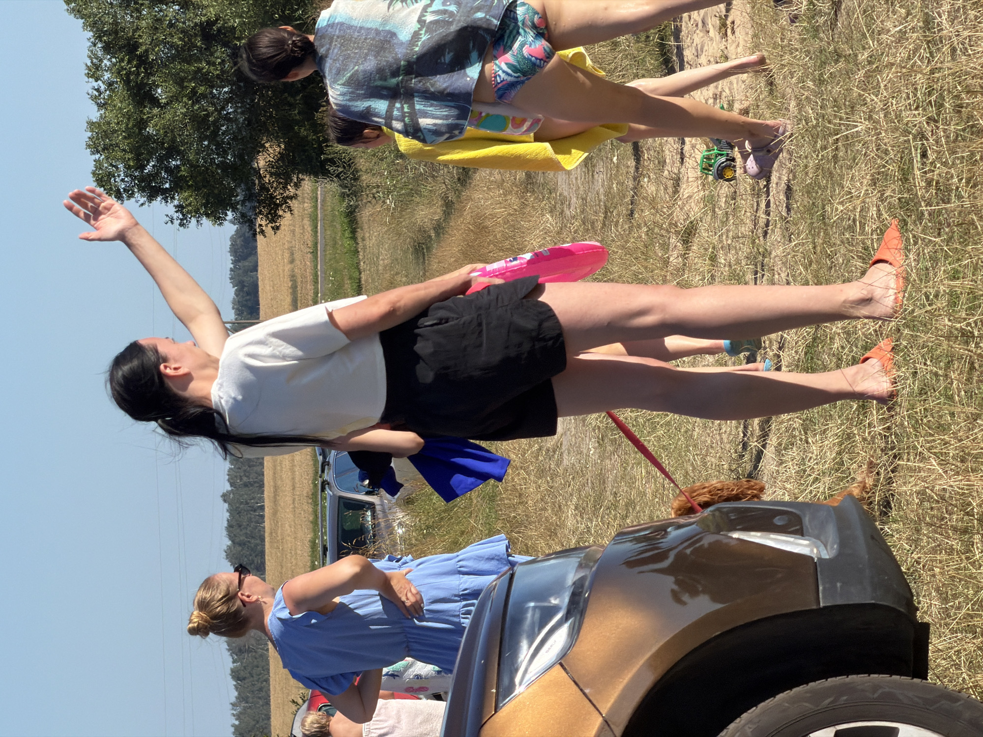 In Валевачскі сельскі Савет, Belarus, on a sunny day, a group of people are gathered in an open, grassy area with a few cars parked nearby. The scene appears to be a casual outdoor event, possibly a picnic or a gathering near a body of water, given the presence of swim attire.
In the foreground, a golden-brown car is partially visible on the left side of the frame. A small, shaggy, brown dog is tethered to a red leash, which is connected to the car and extends towards the right side of the image. The dog is lying down on the dry grass.
Standing near the car is a person wearing orange slip-on shoes and black shorts. They are holding a pink inflatable toy. Behind them, a woman in a blue dress is looking to her left, with her hand on her hip. Another person, wearing a white t-shirt, is turned away from the camera, with their right arm raised as if waving or gesturing.
Further to the right, a young person in a yellow towel is visible, and behind them, another individual is wearing a patterned swimsuit and a colorful scarf draped over their shoulders. There are other people in the background, partially obscured by the foreground subjects and the cars. The environment is characterized by dry, sun-baked grass, a few scattered trees in the middle ground, and a distant tree line against a clear blue sky. A small green toy tractor is lying on the ground near the feet of one of the individuals in swim attire.