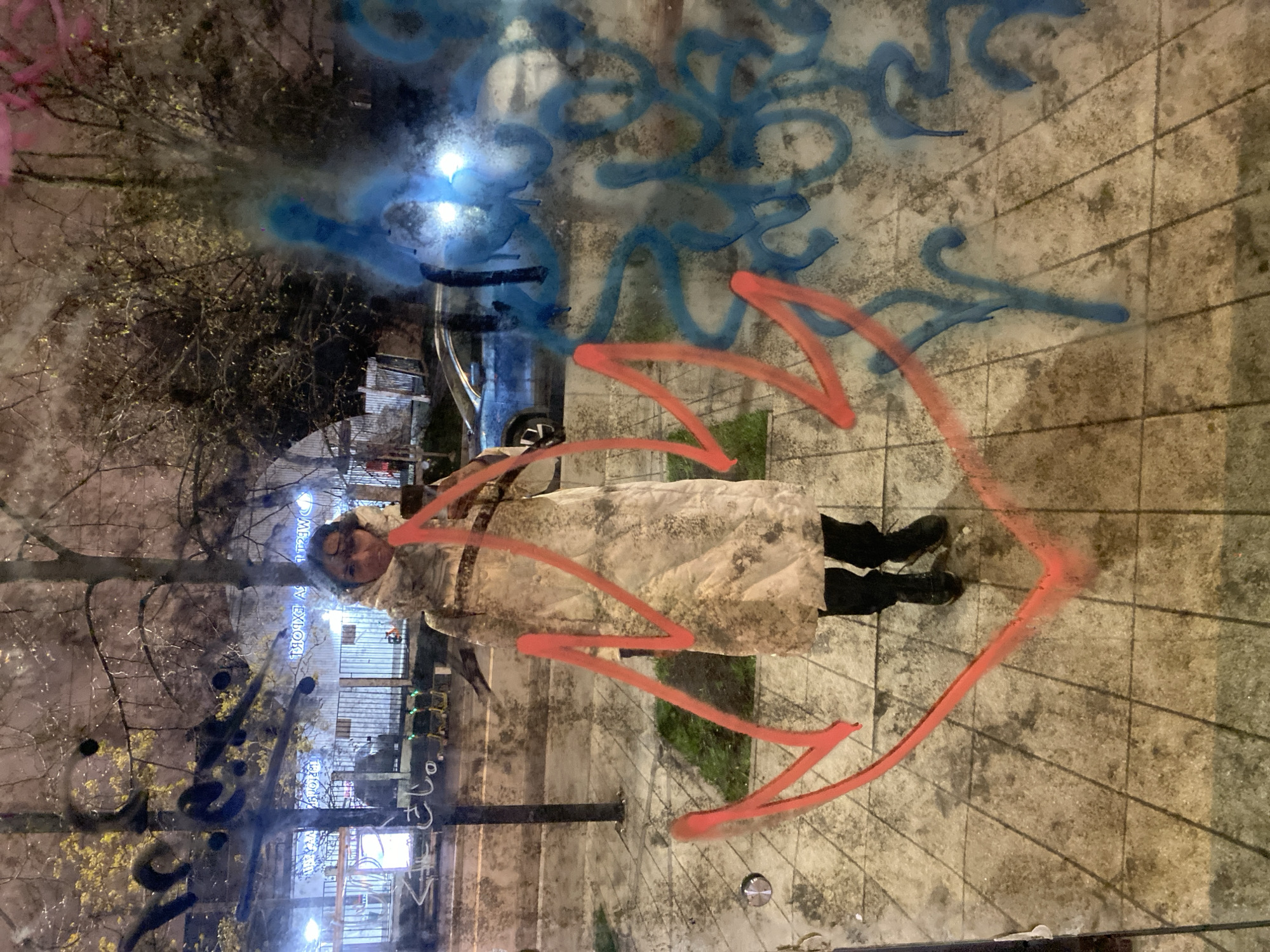 A woman in a light-colored coat and black boots stands facing the camera, seemingly posed in front of a glass surface with graffiti. The graffiti consists of blue and red paint, with a red arrow-like shape surrounding the woman. The background shows an urban environment at night, with trees, streetlights, buildings, and parked cars visible through the glass and graffiti. The scene appears to be in Moscow, Russia, based on visible signage in the reflection.