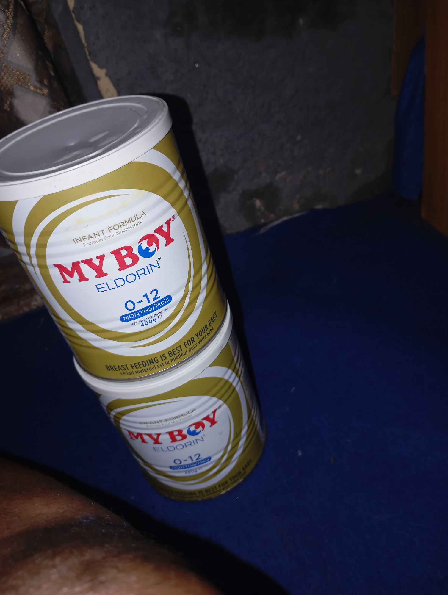 The image shows two stacked cans of "MY BOY ELDORIN" infant formula. The top can is mostly in focus, displaying the brand name, "MY BOY," in red letters with a blue and white logo above the "O". Below that, "ELDORIN" is written in blue letters. The can also indicates "INFANT FORMULA," "Formule Pour Nourrissons," "0-12 MONTHS/Mois," and "NET WEIGHT/Poids net 400g e". There is also text in smaller font at the bottom of the can that reads "BREAST FEEDING IS BEST FOR YOUR BABY Le lait maternel est le meilleur pour votre bébé". The second can is partially visible beneath the top can and appears to have the same branding. Both cans are predominantly white with gold circular accents. The cans are placed on a dark blue surface, possibly fabric. The background is dark and out of focus, with hints of texture that suggest a wall or draped material. The lighting is dim, with a strong light source coming from the right side of the image. The overall scene is indoor and appears to be in a residential setting.