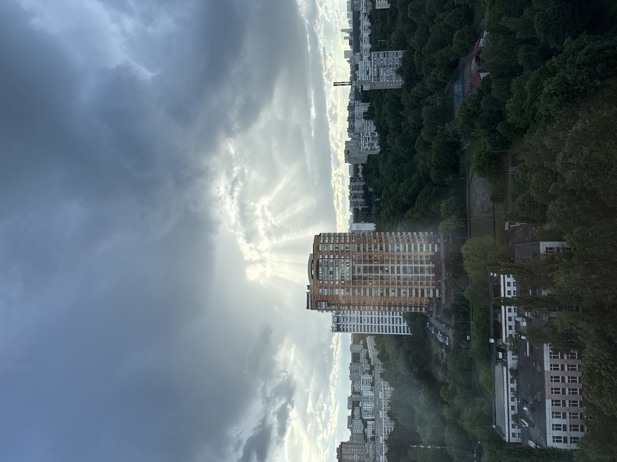 The image depicts a panoramic view of a residential area in Moscow, Russia, under a dramatic, cloudy sky. The sun is breaking through the clouds, casting visible rays of light onto the cityscape. In the foreground, there are several low-rise buildings with visible windows and rooftops, surrounded by dense green trees. Further in the background, a cluster of mid-rise apartment buildings of varying architectural styles and colors are visible, with a particularly prominent orange-brick high-rise apartment building standing out in the mid-ground. Beyond these, a more distant skyline of modern, taller buildings can be seen, hinting at the larger urban expanse. A patch of parkland or a sports complex with tennis courts is visible amidst the trees in the mid-ground. The overall impression is one of a densely populated urban environment interspersed with significant green spaces, under a dynamic and atmospheric sky.