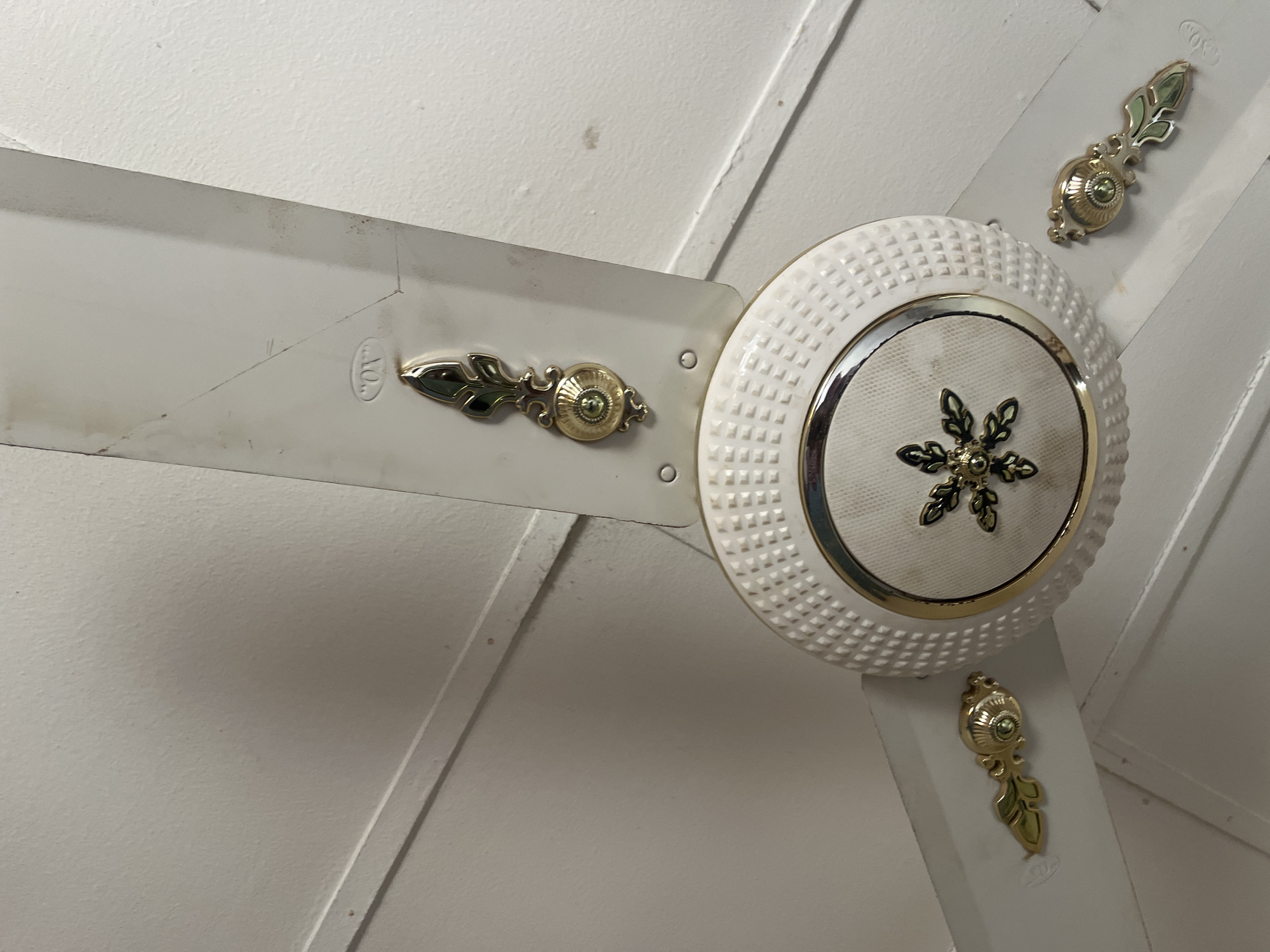 The image shows a white ceiling fan with decorative gold accents. The fan has three blades, each with a gold ornamental piece attached near the end. The central housing of the fan is white and textured, with a gold rim and a decorative snowflake-like design in the center. The fan is mounted on a white ceiling, which appears to be made of panels. The overall impression is one of a functional household item with some decorative embellishments. The location is described as Jalingo, Nigeria, though this information is not visually verifiable from the image itself.