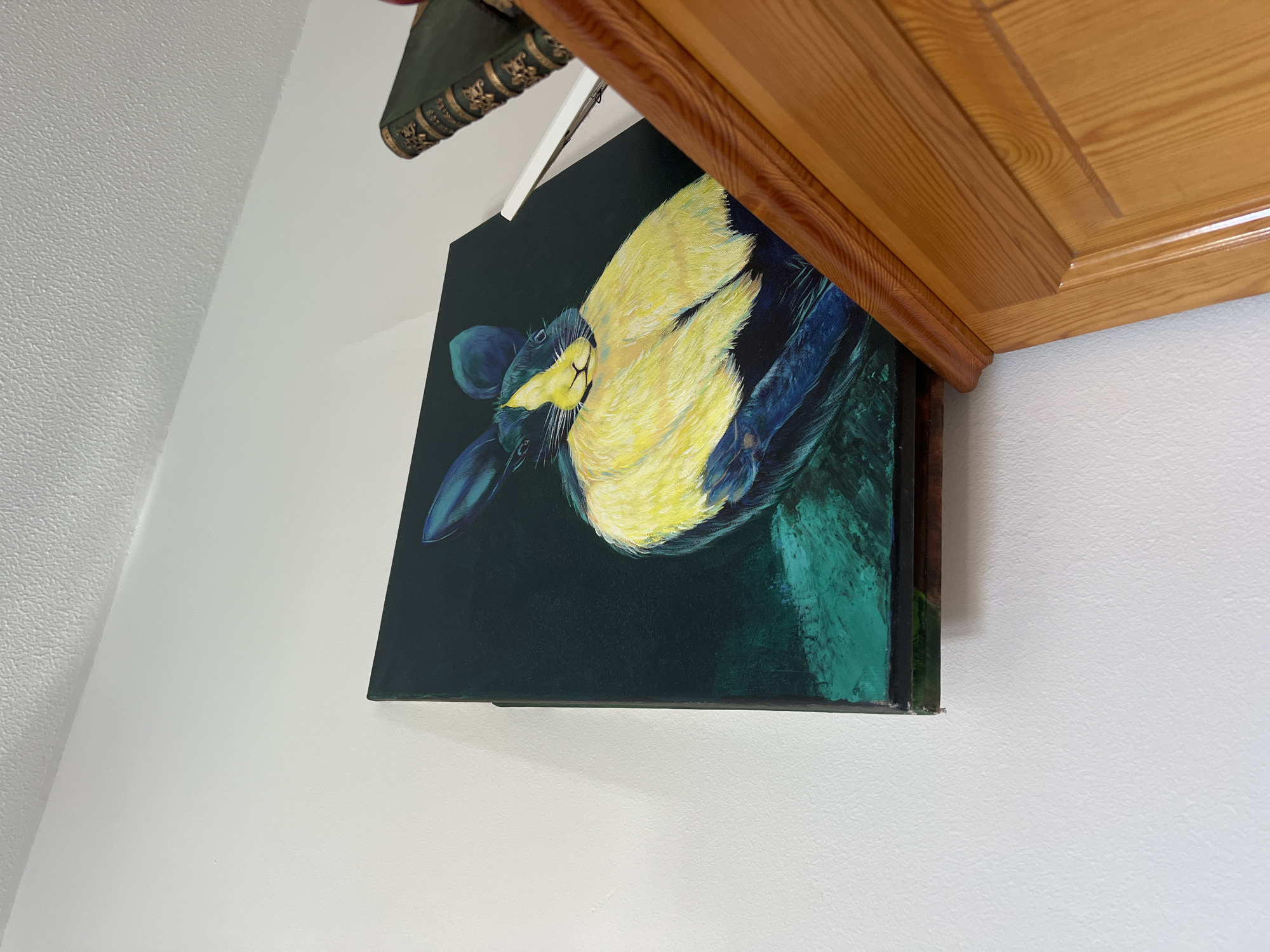 A painting of a rabbit is displayed on a white wall in Oroklini, Cyprus. The painting is in portrait orientation and depicts a rabbit with dark blue ears and a yellow and white chest. The background of the painting is dark green. To the right of the painting, on a wooden surface, are two antique-looking books. The top book is green with gold lettering and ornate detailing, while the bottom book is not fully visible. The lighting suggests an indoor setting, likely a room with natural light coming from an unseen source.