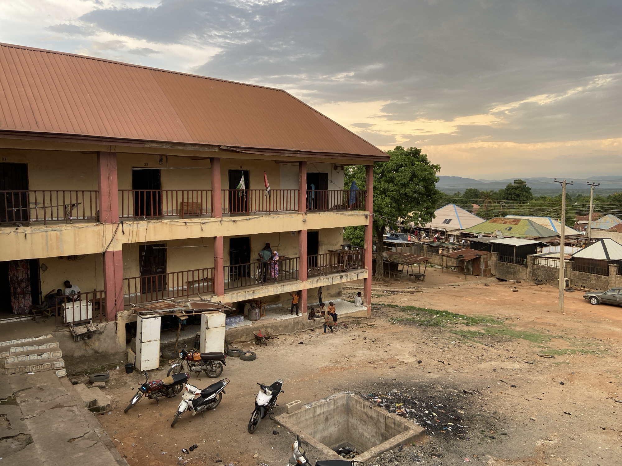 The image depicts a scene in Tudun Wada Bibinu, Nigeria. A two-story building with a brown roof and beige walls dominates the left side. The building features balconies with wooden railings. People are visible on the balconies and in the ground floor area. Three motorcycles are parked in front of the building. Several discarded refrigerators stand near the building's base. In the foreground is a concrete structure filled with debris, suggesting a drainage or waste disposal point. The ground is uneven, mostly dirt and gravel.
In the background, a cluster of smaller buildings with various roof types (metal sheets, tiles) is visible. A few trees are scattered throughout. Power lines are strung between utility poles. A light-colored sedan is parked on the right edge. The sky is overcast, suggesting early morning or late afternoon lighting. Several individuals are on the balconies of the building as well as the courtyard, appearing to be conversing with each other. Some children are sitting or playing on the ground in the building’s courtyard.