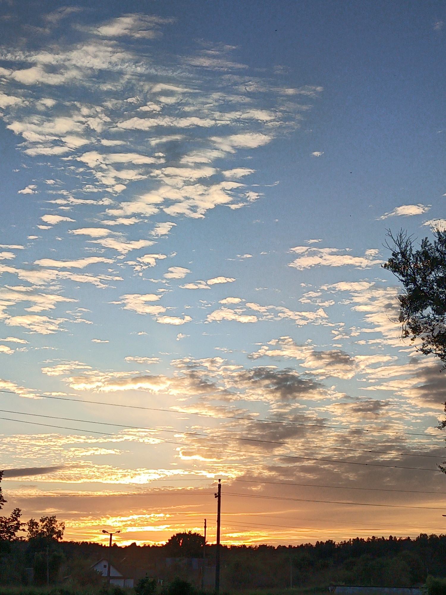 The image depicts a sunset over a rural landscape in Paryechcha, Belarus. The sky is filled with a mix of blue and orange hues, with scattered white and gray clouds. The sun is low on the horizon, casting a warm glow. Silhouetted against the sky are a line of trees, power lines, and utility poles. A small house with a white roof is visible among the trees in the lower left portion of the image. The overall scene conveys a sense of peace and tranquility.