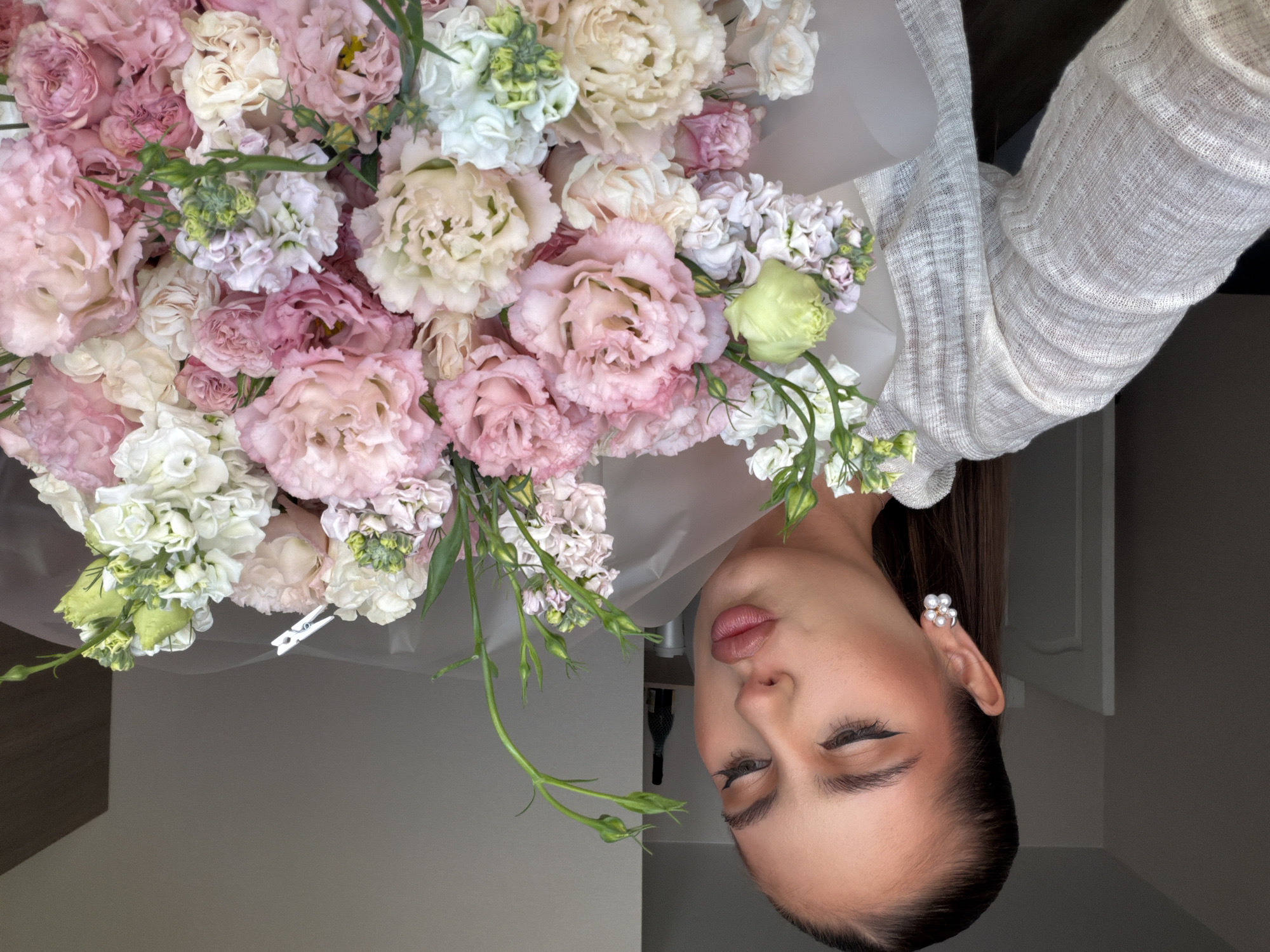 A fair-skinned woman with dark hair is holding a large bouquet of pink, cream, and white flowers. She is wearing a light-colored, knit sweater. A white pearl earring is visible in her left ear. She is facing forward with her eyes closed. The background is a neutral gray, partially obscured by the bouquet. A dark bottle is partially visible behind her head. The location is not discernible from the image.