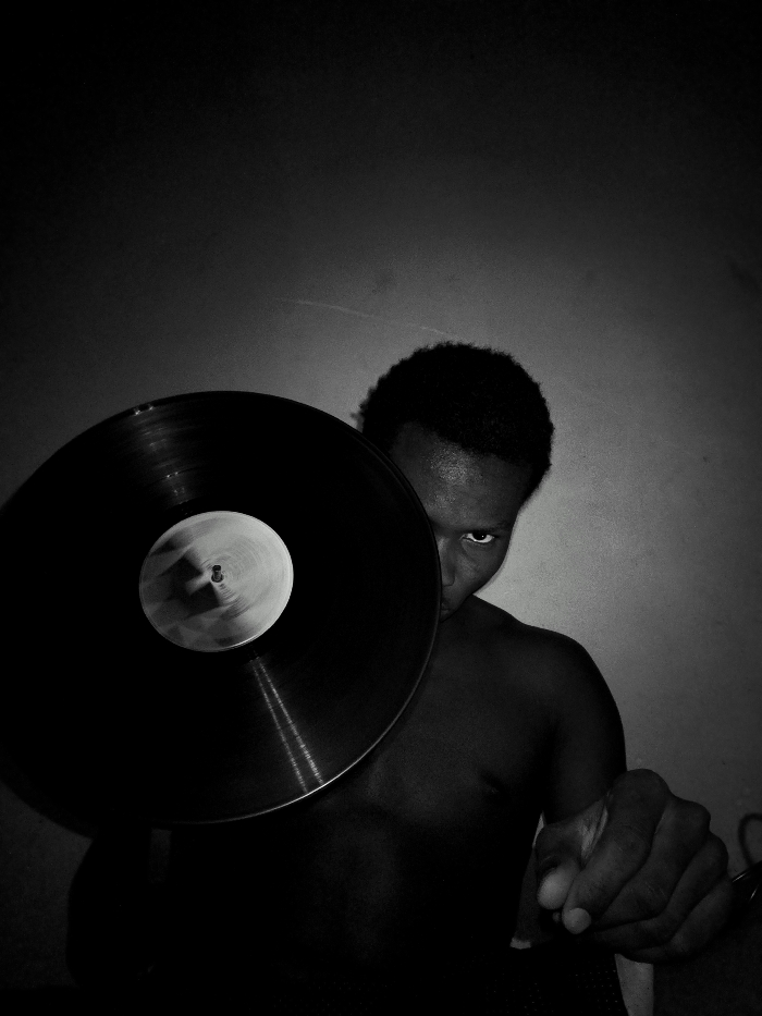 A person with short hair peers from behind a vinyl record, partially obscuring their face. The subject is shirtless, with their right hand extended towards the viewer, forming a fist. The image is in grayscale, with a dark background highlighting the contrast between the record's surface and its label. The overall composition creates a sense of mystery and intensity.