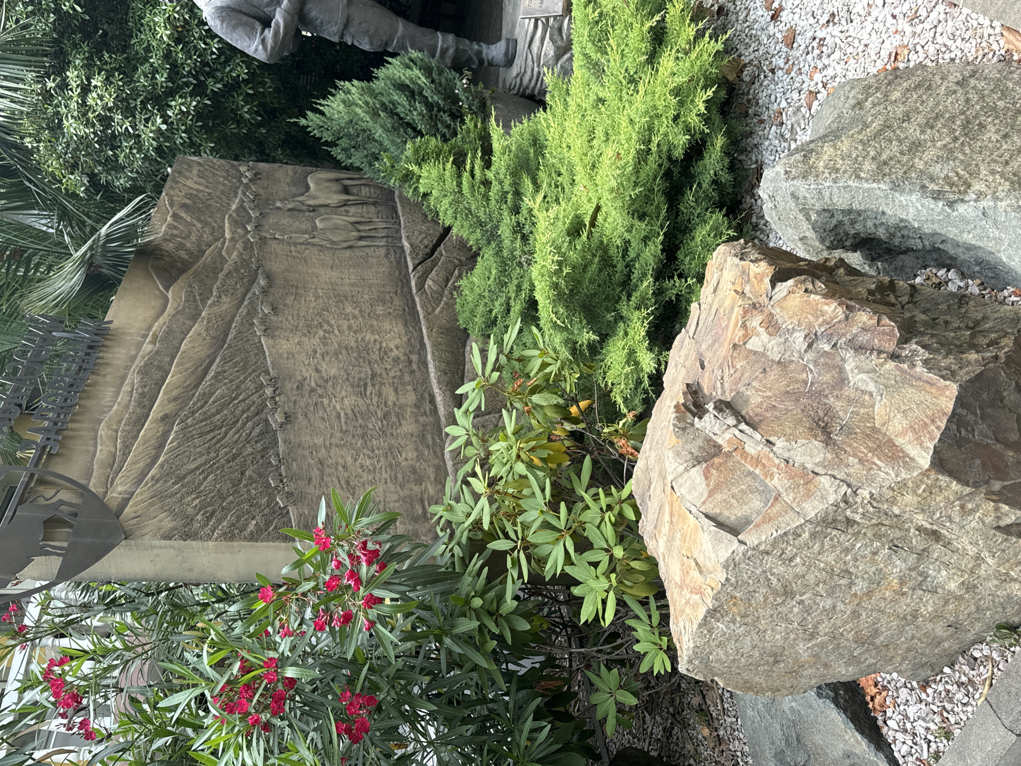 The image depicts an outdoor monument with a carved stone relief of a mountain landscape featuring animals. In the foreground, there are large rocks and various green plants, including flowering bushes with red blossoms and coniferous shrubs. To the right, the lower portion of a bronze statue of a person in uniform is visible. The monument appears to be located in a landscaped area with gravel ground cover. This scene is located in the Arboretum Botanical Garden in Sochi, Russia, specifically at the monument dedicated to the mountain hunters.