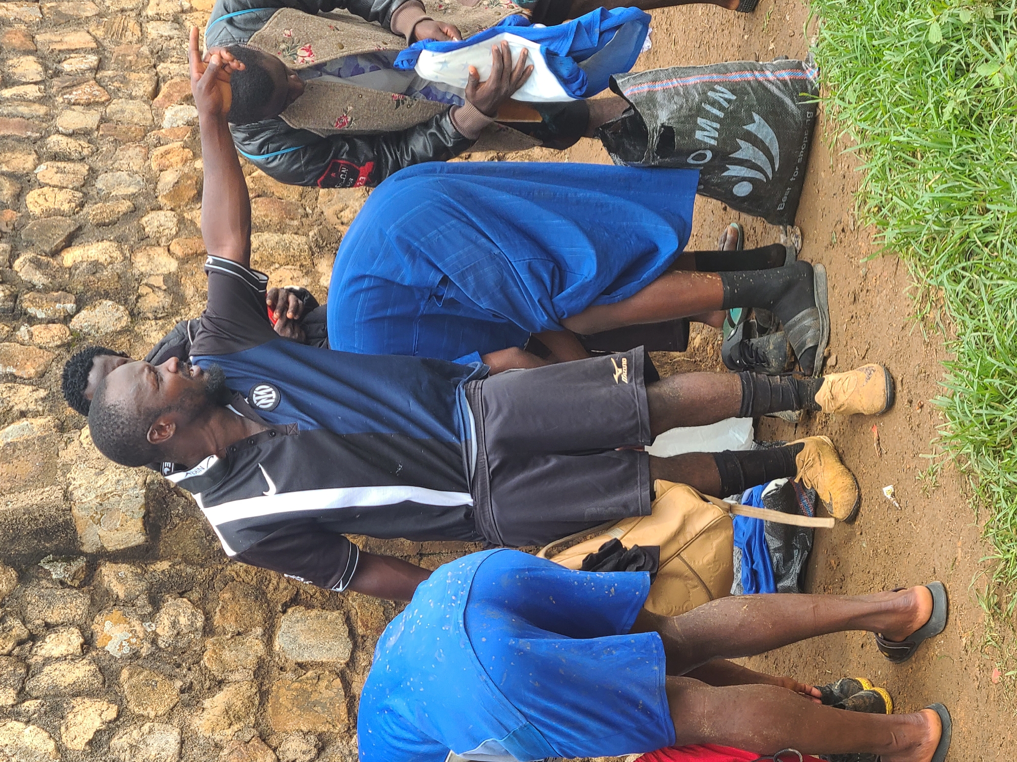 The image depicts a group of at least four individuals outdoors, situated near a wall constructed from irregularly shaped stones. The location appears to be urban, with grassy areas and a dirt or gravel ground surface visible. One individual is wearing a black and blue athletic shirt with the Inter Milan logo. Another individual wears a blue skirt. A person on the far left of the image is holding a cloth. The individuals are close together, with some appearing to be kneeling or sitting. Additional details include the presence of a bag with the word "OMIN" printed on it, along with various items of clothing and footwear scattered nearby. The overall impression is one of a gathering or possibly a makeshift living situation.