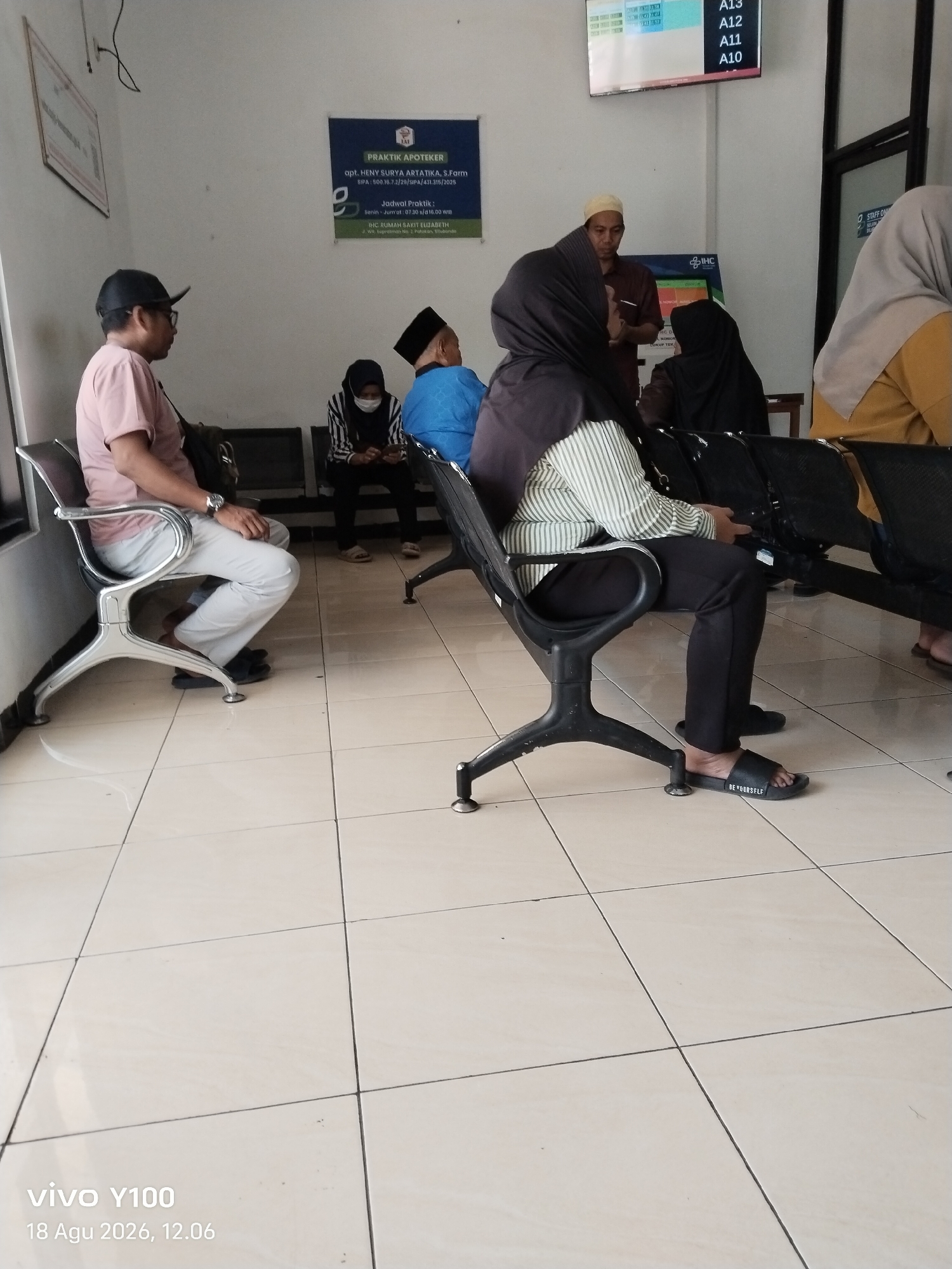 The image depicts the waiting area of an Indonesian health facility, likely a clinic or hospital, in Patokan, Indonesia. Several individuals are seated on black waiting chairs, facing a tiled floor. In the foreground, a man wearing a pink t-shirt and a black cap is seated. To his right, a person wearing a striped long-sleeved shirt and black pants is also seated, with their feet visible wearing black slides that say "BE YOURSELF". Further back, other individuals are present, including a woman in a hijab wearing a mask and looking at her phone, and a man in a maroon shirt and a white cap who appears to be speaking to someone. A woman in a light brown hijab is visible in the far right of the frame. The environment is well-lit and appears to be indoors, with white walls. A sign with "PRAKTIK APOTEKER" and a digital display board showing "A13", "A12", "A11", and "A10" are visible on the wall. The date "18 Agu 2026, 12:06" is also present in the lower left corner, indicating a future date.