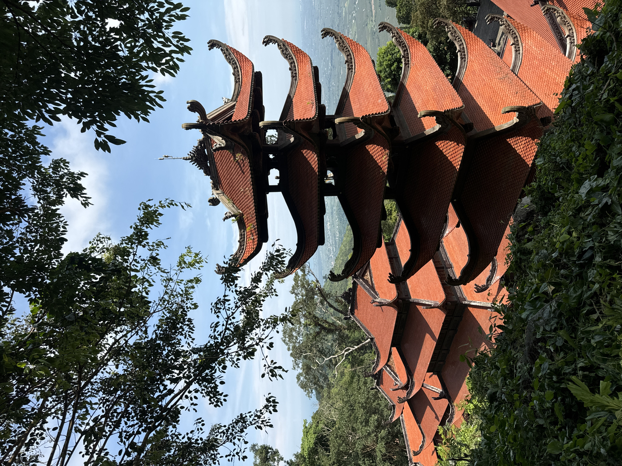 In Thuận Nam, Vietnam, the image captures a multi-tiered pagoda with a distinctive red-tiled roof. The pagoda is set against a backdrop of lush green foliage and trees, with a hazy landscape of buildings and terrain visible in the distance. The sky is a clear blue with scattered white clouds. The pagoda's roofs are intricately designed with curved edges and decorative elements, suggesting traditional architectural style. The foreground is dominated by dense green vegetation, partially obscuring the lower sections of the pagoda. The lighting suggests it is daytime, with sunlight casting shadows on the tiled roofs. No people or specific actions are depicted; the scene focuses on the architectural structure and its natural surroundings.
