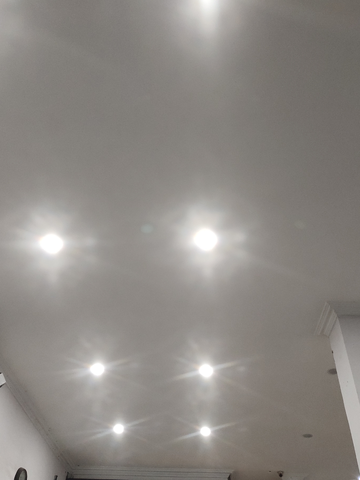 The image is a low-angle, full shot of a white ceiling with recessed lighting fixtures. There are six circular lights visible, each emitting a bright glow with starburst-like reflections. The ceiling appears to be a standard drywall or plastered surface, with some minor imperfections or dust specks visible upon close inspection. In the lower right corner, a portion of a white wall with decorative molding is visible, suggesting an indoor environment. No people or specific identifiable objects are present in the frame besides the lighting fixtures and architectural elements. The scene is devoid of any context that would place it in a specific city or location, including Lhoksukon, Indonesia.