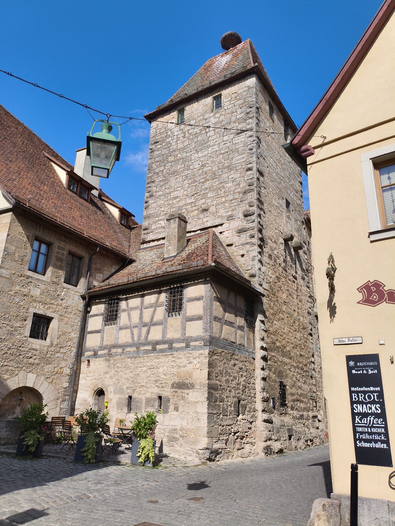 The image shows a view of a narrow cobblestone street in Rothenburg ob der Tauber, Germany. On the right side of the street, a light yellow building with a dark red roof trim has a black sign advertising "BROTHAUS Brot & Zeit" which offers "HOLZOFENBROT BROT, SNACKS, Kaffee, TORTEN & KUCHEN, FRÜHSTÜCK, SCHNEEBALLEN". The sign also indicates "Alter Keller" above it. To the left of this building is a tall, ancient stone tower, possibly a part of the city's fortifications, with a tiled roof topped by a bird's nest. Attached to the tower is a smaller, timber-framed structure with a red tile roof. In front of the tower's timber-framed section, there are several potted plants and outdoor seating with tables and chairs. A vintage-style street lamp hangs from a wire spanning the street. The sky is clear and blue.