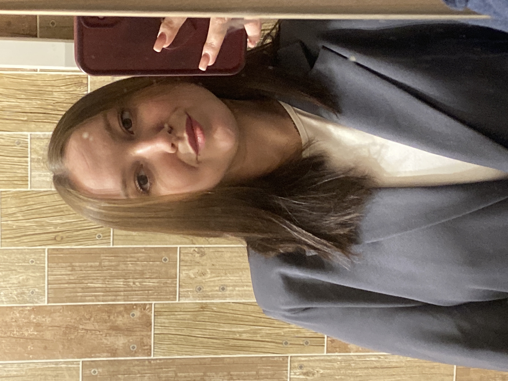 A woman is taking a selfie in front of a wall covered in wood-patterned tiles. She is holding a maroon-colored smartphone and wearing a gray blazer over a white top. Her hair is shoulder-length and light brown. The location appears to be indoors, possibly a bathroom or a room with similar tiling. Chaikovsky, Russia, is not discernible from the image.