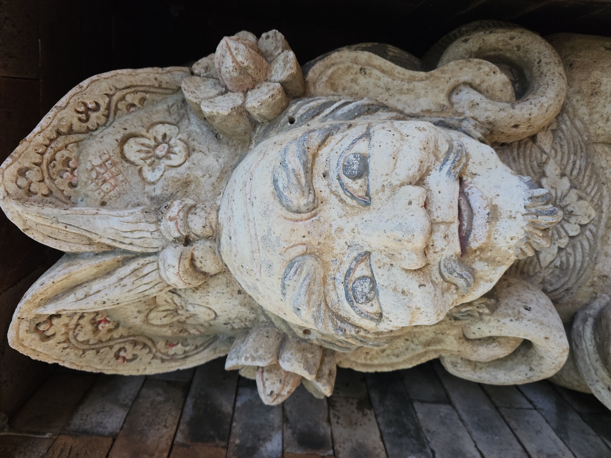 This is a close-up, eye-level photograph of a weathered, stone statue of a grotesque figure's head. The statue is light beige and appears to be made of concrete or a similar material, with some areas showing signs of age and erosion. The figure has a large, bulbous head with a wrinkled forehead and prominent, wide-open eyes outlined in a faded blue. Its nose is broad and flat, and its mouth is open in a grimace, revealing a few teeth and a protruding tongue. A thin, stylized mustache and a pointed beard adorn its chin. The figure wears an elaborate headdress featuring floral motifs, decorative swirling patterns, and what appear to be stylized feathers or hair extensions framing its face. The statue is positioned against a dark background, with a textured, brick-like surface visible to the left of the frame. The lighting is harsh, casting shadows that accentuate the statue's weathered features. This statue is located in Denpasar, Indonesia, likely as a decorative element in a temple or traditional Balinese architectural setting.