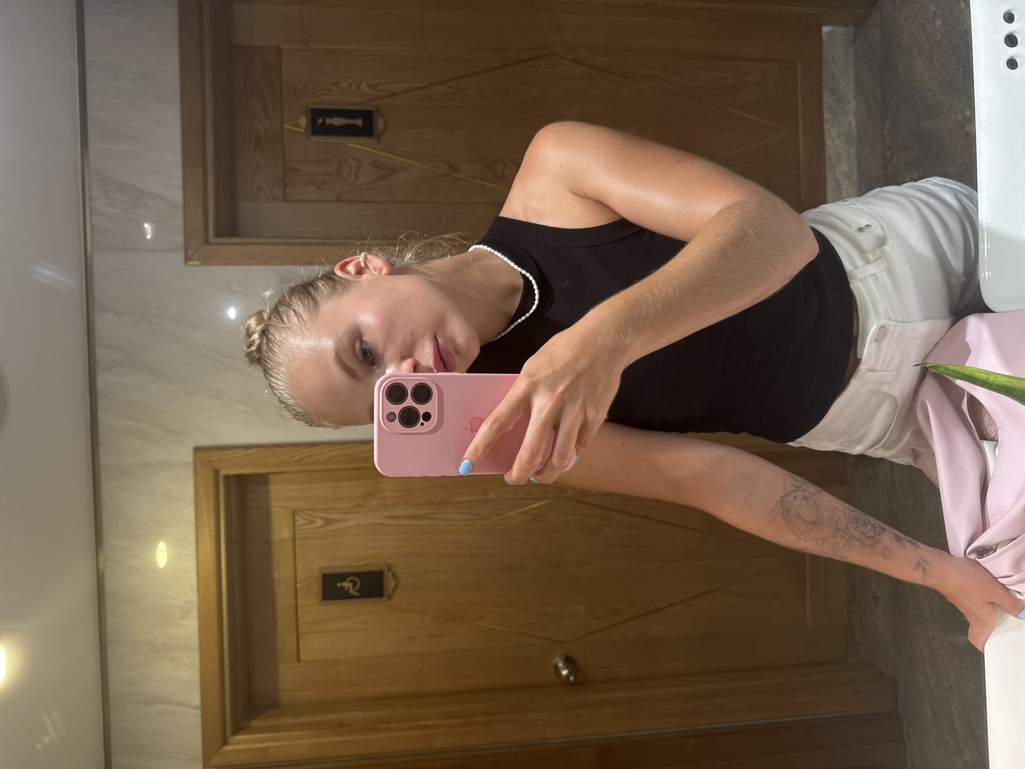 A young Caucasian woman with blonde hair pulled up in a bun takes a selfie in a mirror. She is wearing a black tank top, white shorts, and a pearl necklace. Her left arm has a tattoo of a dragon and flowers. Her fingernails are painted light blue. She is holding a pink smartphone in her right hand. The background appears to be a public restroom with two wooden doors, one with a disabled access symbol and the other with a female symbol. The walls are tiled with a marble pattern. The woman is standing in front of a white sink.