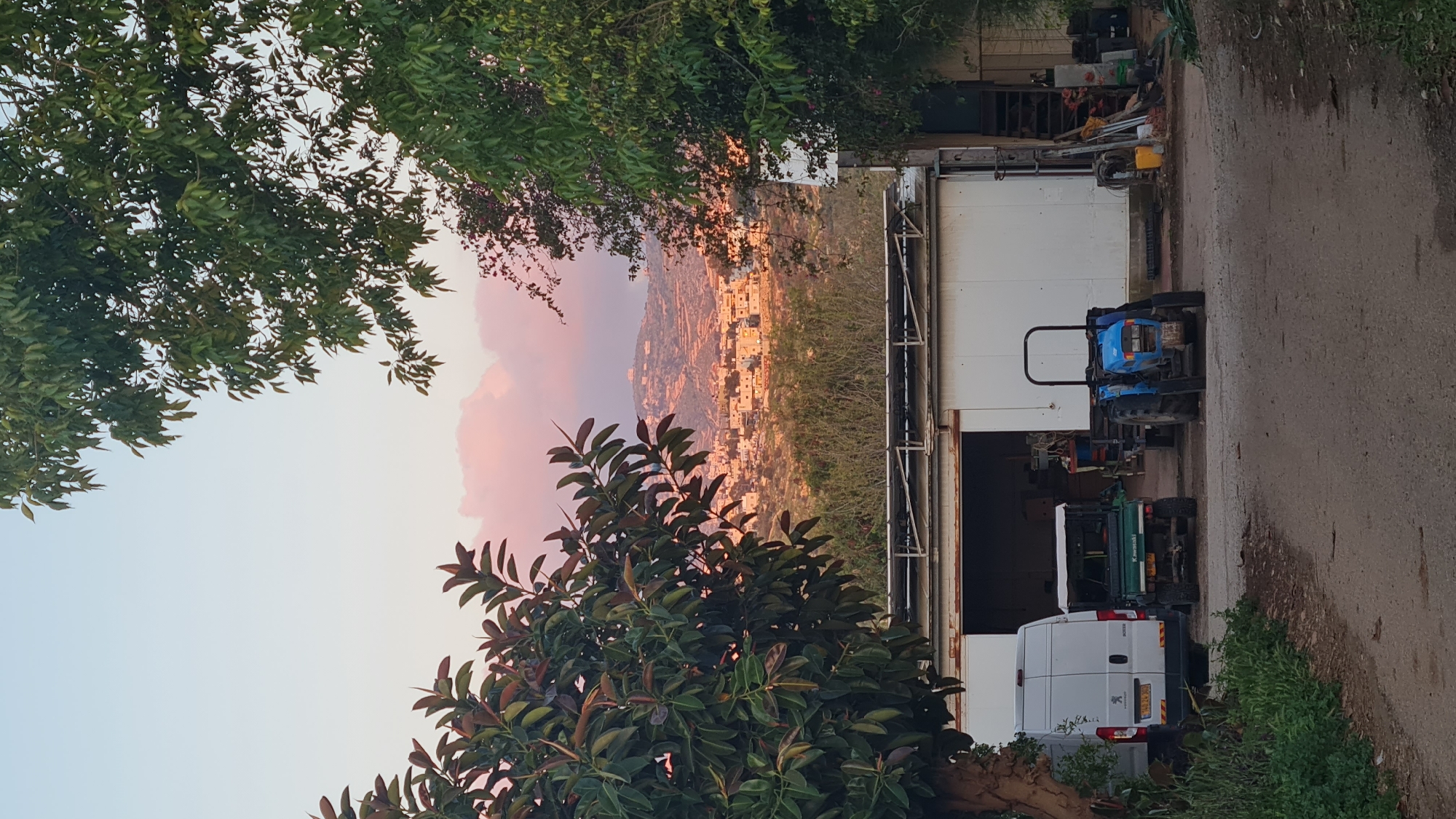 The image depicts a view in Lachish Regional Council, Israel, at sunset. In the background, a pink-hued sky illuminates a distant city perched on a hillside. In the foreground, foliage frames the sky, with a white building extending to the right. A white Peugeot van is parked in front of the building, followed by a green Kawasaki and a blue tractor. A dirt road runs along the right side of the image, with vegetation along its edge. A collection of metal objects sits adjacent to the white building.