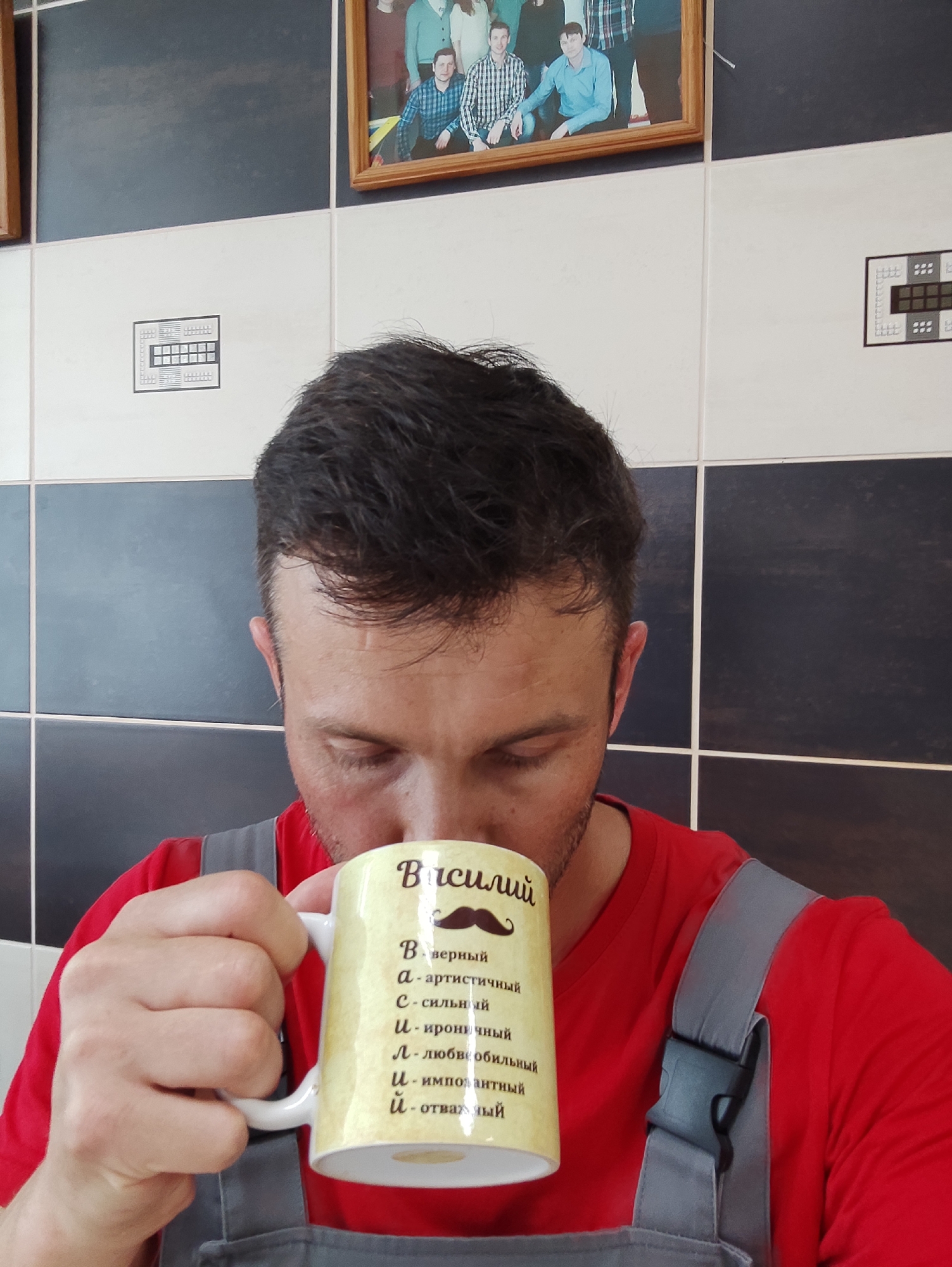 An individual is depicted from the upper chest upwards, consuming a beverage from a mug in an indoor setting. The subject, a male, has short, dark hair and visible stubble. He wears a red t-shirt beneath gray overalls, with a black plastic buckle visible on the right shoulder strap. His eyes are downcast as he holds a yellow/cream-colored mug with a white handle in his right hand, positioned at his mouth.
The mug bears Russian text: "Василий" (Vasily/Basil) above a stylized mustache icon. Below this, an acrostic poem describes traits associated with the name:
* В - верный (faithful)
* а - артистичный (artistic)
* с - сильный (strong)
* и - ироничный (ironic)
* л - любвеобильный (affectionate)
* и - импозантный (imposing)
* й - отважный (brave)
The background consists of a tiled wall featuring alternating black/dark gray and white/light gray square tiles arranged in a checkerboard pattern, separated by white grout lines. Mounted on the wall are two small framed diagrams or schematics, each displaying grids and various symbols. One is visible to the left of the subject's head, and a partial view of another is to the right. Between these, slightly above and to the right of the subject, hangs a framed photograph in a wooden border, depicting a group of approximately six individuals (three men and three women) in casual attire. A partially visible wooden frame is also present on the far left edge of the image. The location is an interior space in Unknown, Unknown.