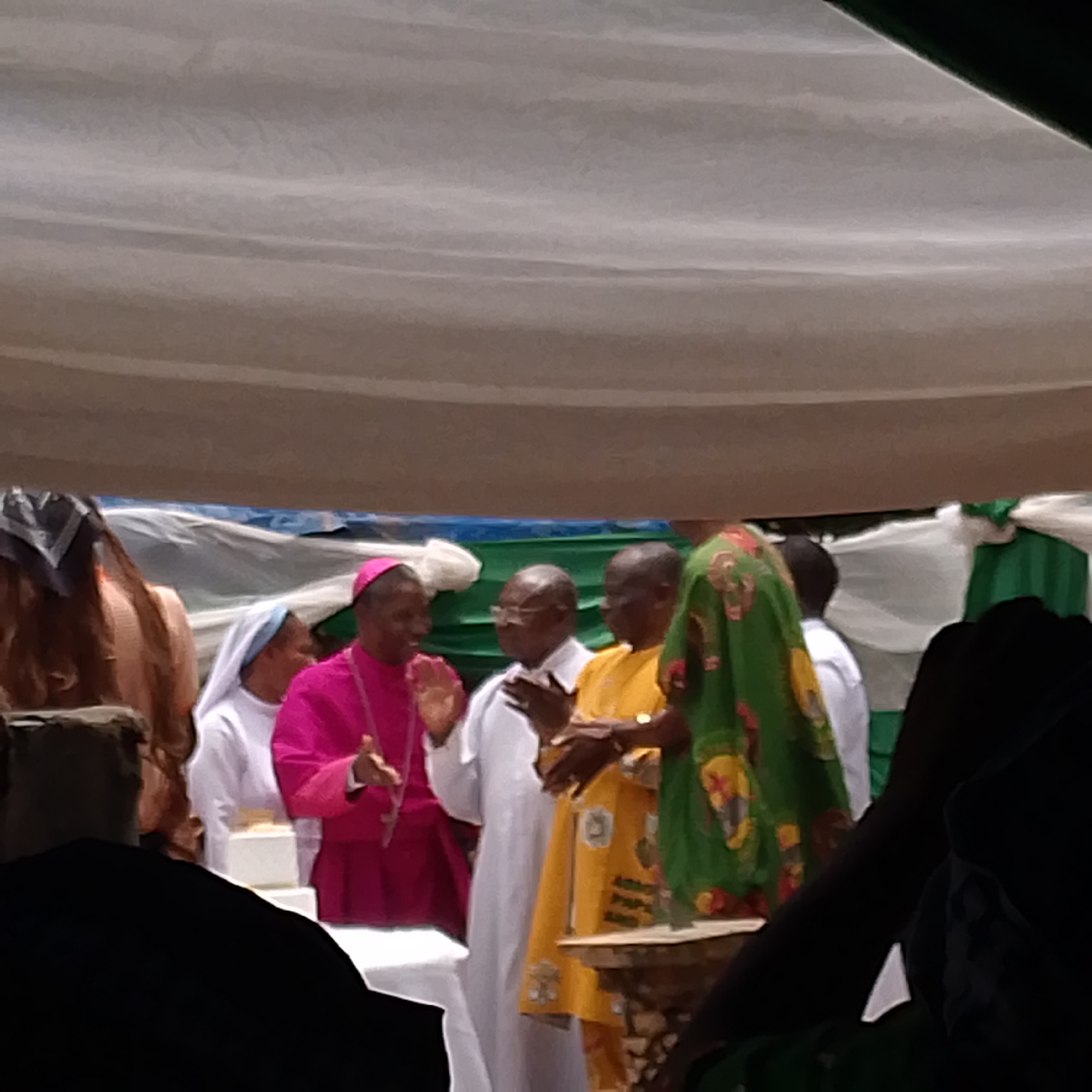 The image shows a group of people gathered outdoors in what appears to be a religious setting in the city of Unknown, Unknown. A bishop in a pink robe is seen speaking with his hands raised. Next to him stands a man in a white robe, and another man in a yellow robe with print on it. To the left, a woman in a nun's habit is visible. A woman wearing a green and patterned head covering stands on the other side of the man in the yellow robe. The scene is partially obscured by shadows and foreground elements. A white canopy stretches overhead, decorated with green and white ribbons.