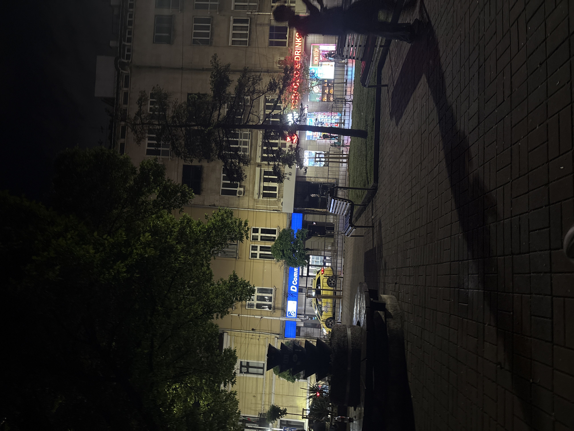 The image shows a night scene in Sofia, Bulgaria. A multi-story building with numerous windows dominates the background. Neon signs advertise a shop selling "TOBACCO & DRINKS" and a "D Com" store. A yellow car is parked in front of the "D Com" store. A tree partially obscures the view of the building. Paved walkway with a bench and a sculpture/monument are in the foreground. A blurry figure is at the top right corner of the image.