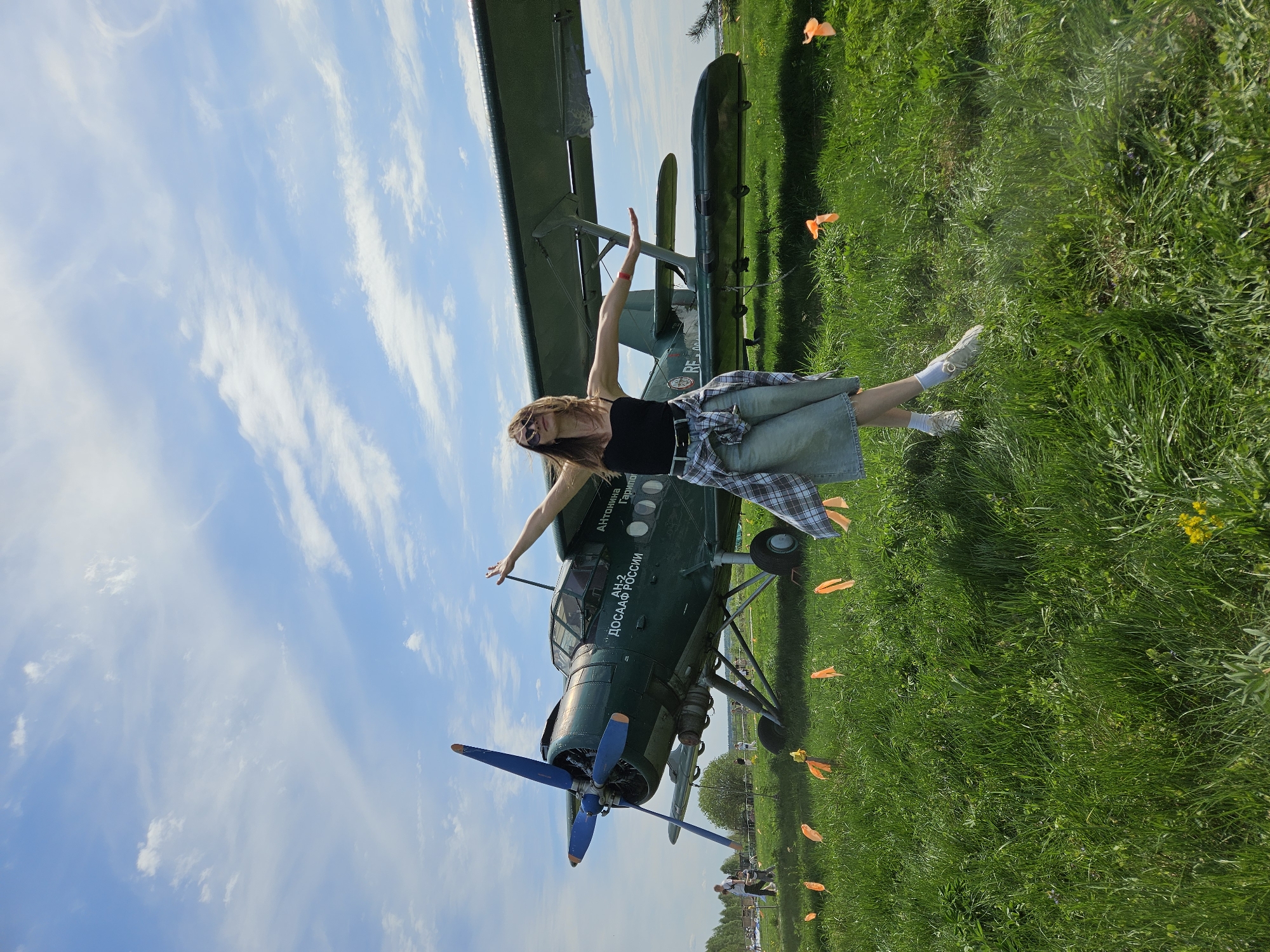 The image shows a fair-skinned young woman standing with her arms outstretched in front of a green biplane. The woman is wearing a black tank top, a green skirt, a flannel shirt tied around her waist, sunglasses, and white sneakers. The biplane is a vintage model, with a blue propeller and the words "AH-2 ДОСААФ РОССИИ" written on the side in Russian. The scene appears to be set on a grassy field with a blue sky and scattered clouds. Small orange markers dot the grass. Based on the plane and the Russian text, the location could be in Russia, but the city of Izhevsk cannot be definitively confirmed from the image alone.
