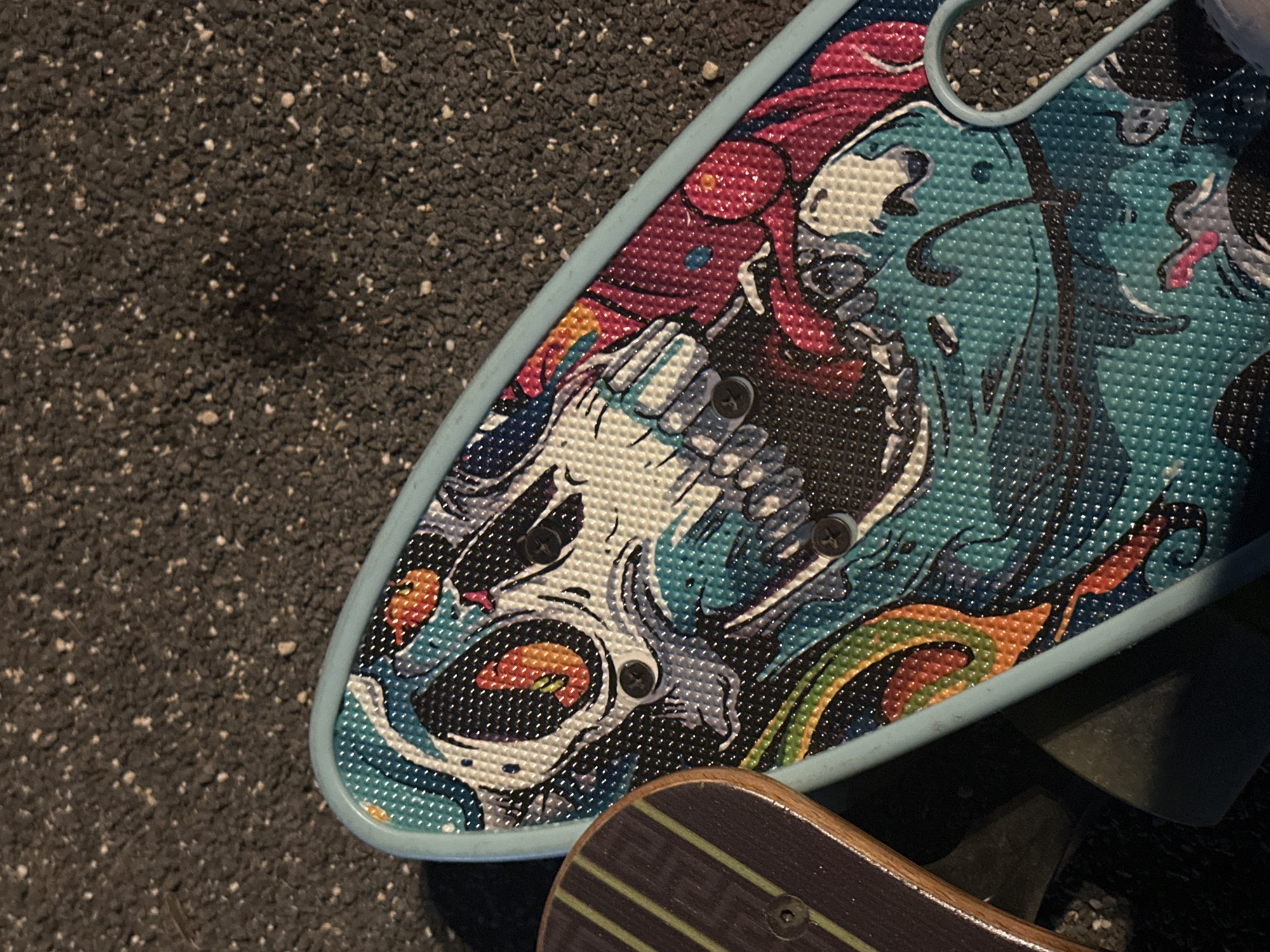 The image shows a close-up of two skateboards on a textured dark gray surface, possibly asphalt or pavement, with scattered small white particles. The skateboard in the foreground has a light blue edge and a colorful graphic design featuring a skull with prominent teeth and eyes, stylized in blues, oranges, whites, and pinks. The skull graphic has a textured, dot-like pattern. Visible on the skateboard are three black screws securing the deck to the trucks. To the left, another skateboard is partially visible. It has a wooden deck with dark brown sides and a pattern of alternating dark green and light green stripes. Only one black screw is visible on this skateboard. The lighting suggests an outdoor setting, possibly during the day, with shadows cast on the surface. The location is not specified in the image.
