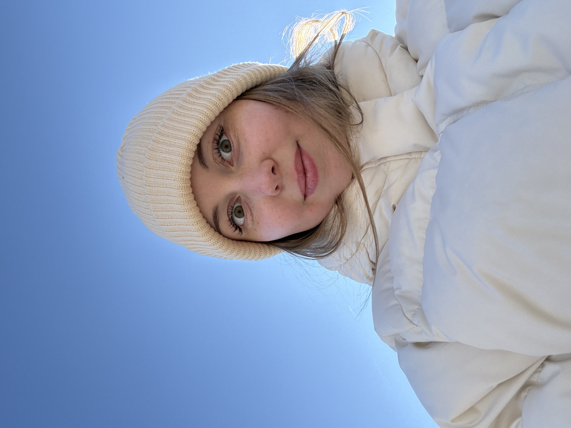 A woman with fair skin and light brown hair is pictured from a low angle, looking down at the camera. She is wearing a cream-colored knit beanie and a puffy, off-white jacket. The backdrop is a clear blue sky.
