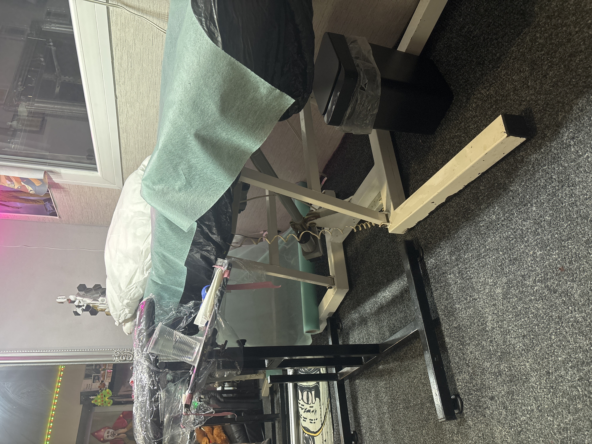 The image shows an examination table in a room with gray carpet. The table is covered with a light green disposable sheet, topped with a white pillow. A black trash bag is draped over one side of the table. The room also contains a metallic-framed vanity mirror with visible reflections of items. The presence of various medical supplies and disposable items scattered around the table suggest this is a medical setting. The location of this photo cannot be determined to be Pleven, Bulgaria.