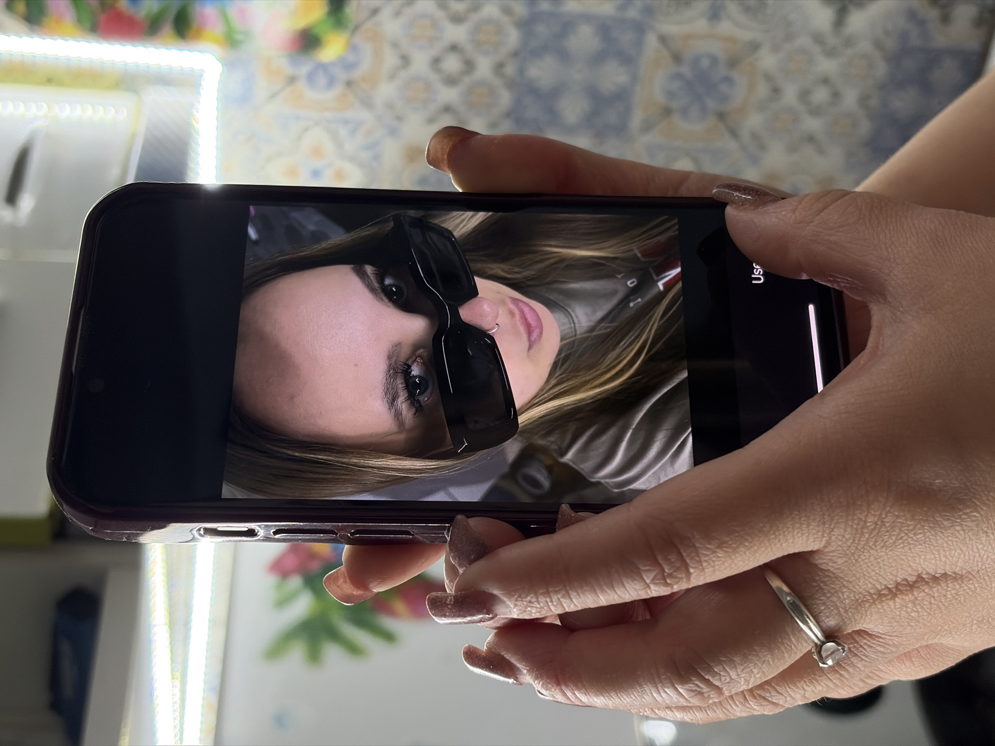 A person holds a smartphone displaying a selfie of a woman wearing sunglasses. The background of the image includes a tiled surface and a light fixture. The subject is inside, the exact location in Port-Katon, Russia is indeterminable.