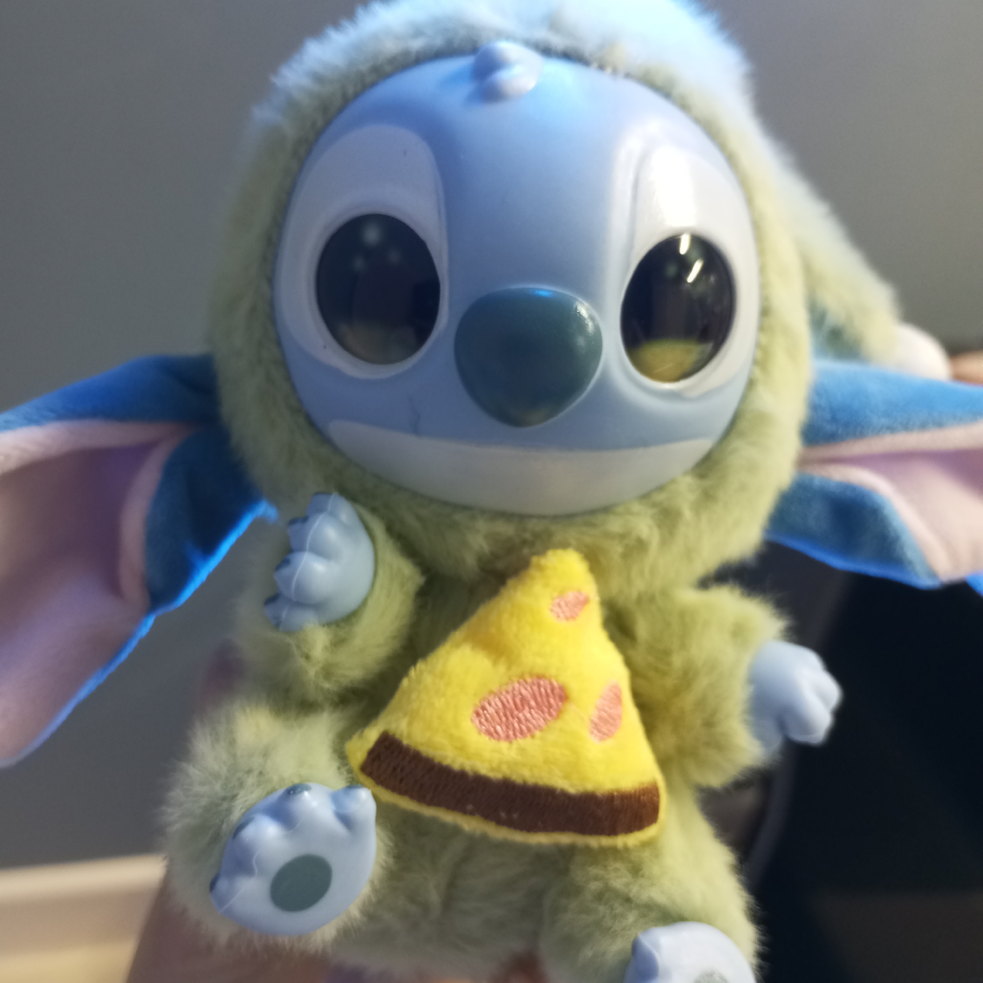 A close-up shot features a toy resembling a blue alien character, possibly Stitch from Disney's Lilo & Stitch, wearing a fluffy, light green hooded costume. The toy has a light blue plastic face with large, dark, reflective eyes and a teal-colored nose. It also has light blue plastic hands and feet. On its back, partially visible, are light blue and pink wings. The toy is holding a soft, yellow, triangular slice of pizza, complete with a brown crust and pink, circular details suggesting pepperoni.
The background is out of focus, showing blurred light gray and darker tones, indicative of an indoor setting. No human figures are visible, but the toy appears to be held by an unseen person. There is no specific activity other than the toy being presented to the camera. No discernible text is visible on the toy or in the background. The image provides no information about the time of day or weather. This media file is associated with the location Pervomaysky, Russia.