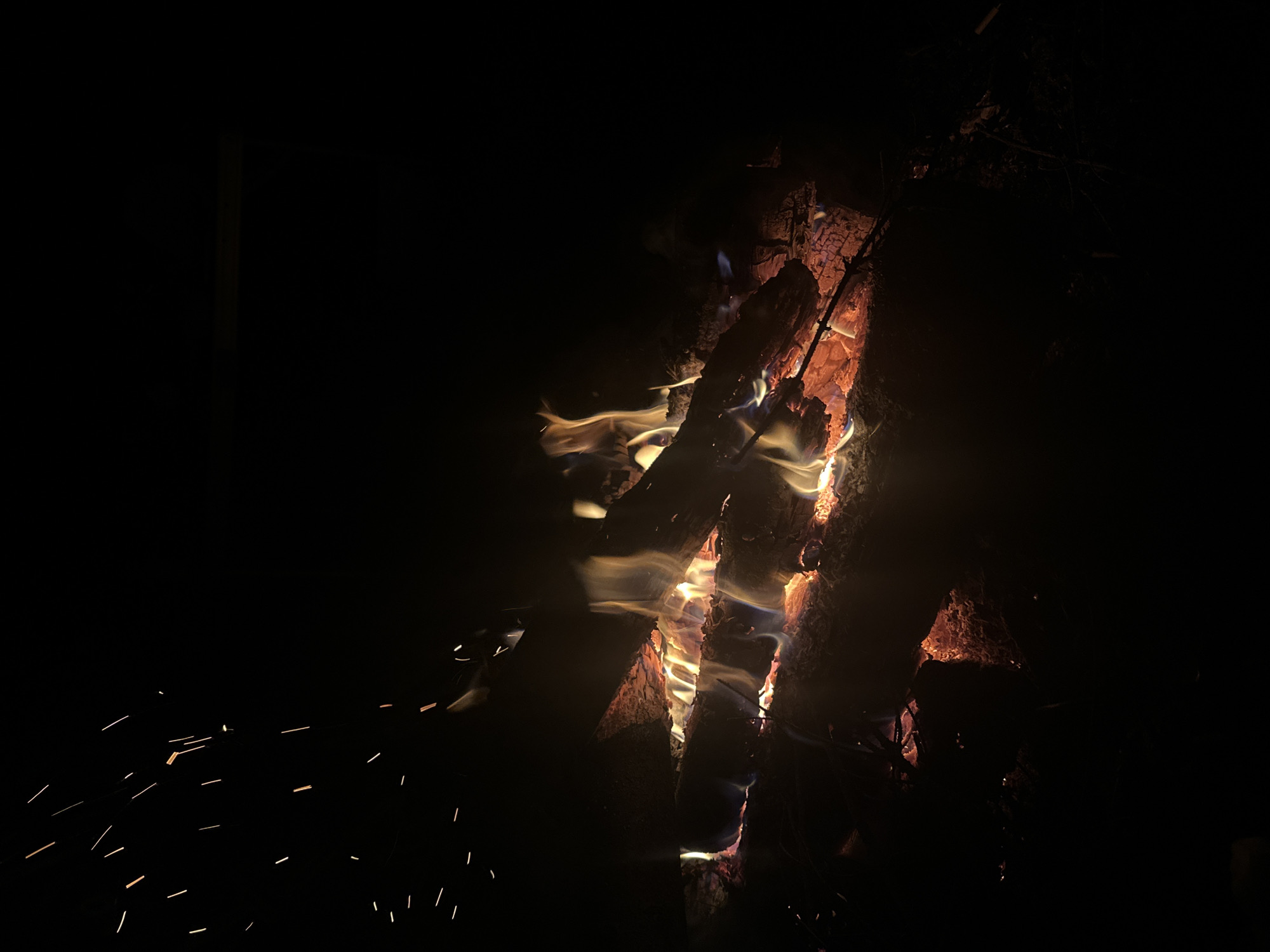 A nighttime scene depicts a close-up view of a bonfire. The fire is built with several pieces of wood arranged vertically. Flames and embers are visible between the logs. Sparks emanate from the fire in the lower-left. The overall ambiance is dark, with the fire providing the primary source of illumination. The location is Папярнянскі сельскі Савет, Belarus. No people are present.