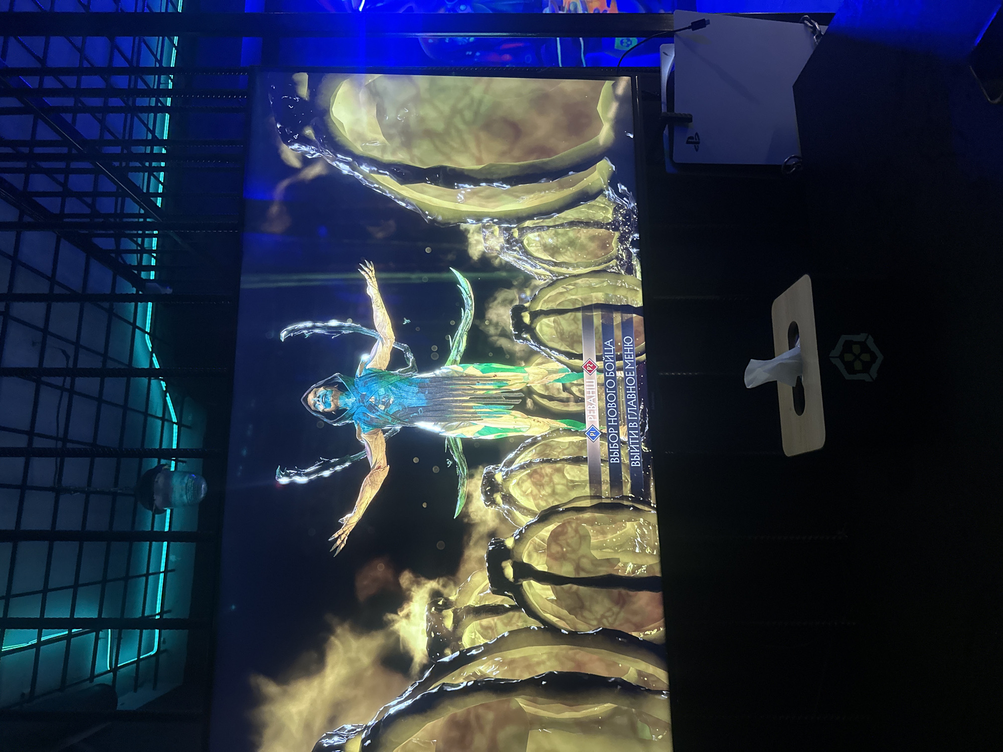 The image shows a television screen displaying a video game menu. The game is likely a fighting game, given the character displayed. The menu is in Russian, with options like "Select New Fighter" and "Exit to Main Menu." The television is situated in a space with a blue-lit metal grid structure on the left side of the image. There's a PlayStation console visible on the right, and also a tissue box. The location (Yerevan, Armenia) is not deducible from the image.