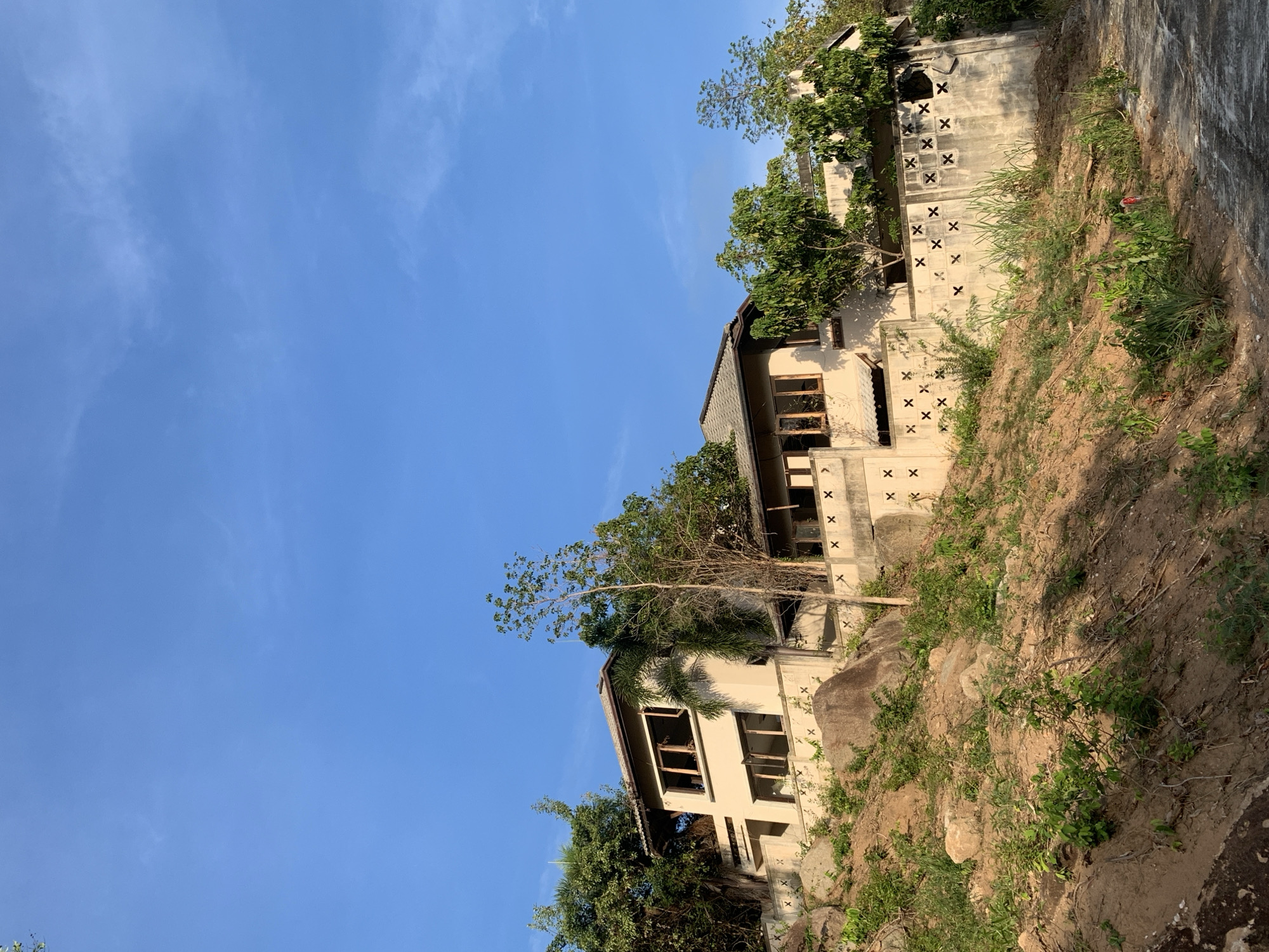 The image captures a multi-story building nestled into a hillside in บ้านบางรักษ์, Thailand. The building is light in color and has a series of windows and decorative cutouts in the wall. It appears to be partially obscured by trees and vegetation, suggesting it may be located in a more natural or undeveloped area. The hillside is covered with dirt, rocks, and sparse plant growth. The sky is a clear blue with minimal cloud cover. The angle of the photo suggests it was taken from a lower vantage point, looking up at the building and sky.