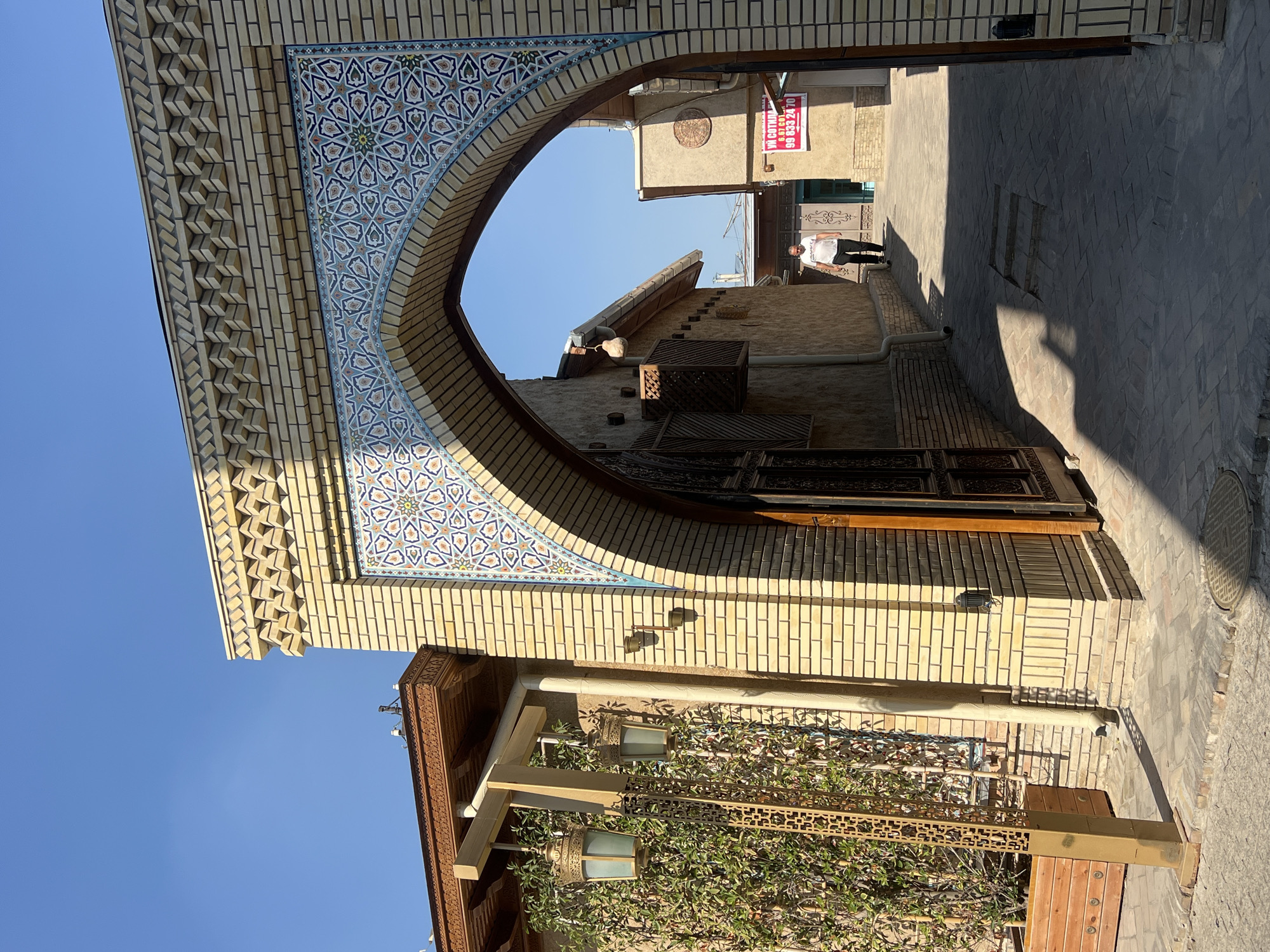 The image shows an arched entryway made of light yellow bricks, decorated with intricate blue and white mosaic tiles. The archway leads to a narrow alleyway paved with cobblestones. Sunlight casts strong shadows across the alley. A man in a white t-shirt and black pants walks away from the camera down the alley. To the left of the archway, there is a building with wooden eaves and two ornate lanterns hanging from wooden beams. Green vines are growing on the side of this building, and a decorative metal screen stands in front of them. To the right of the archway, the alley continues between two buildings with textured beige walls. A red and white sign with Russian text is visible on one of these buildings. The sky is clear and blue. The overall scene depicts a traditional architectural style, possibly in a historical district of Tashkent, Uzbekistan.