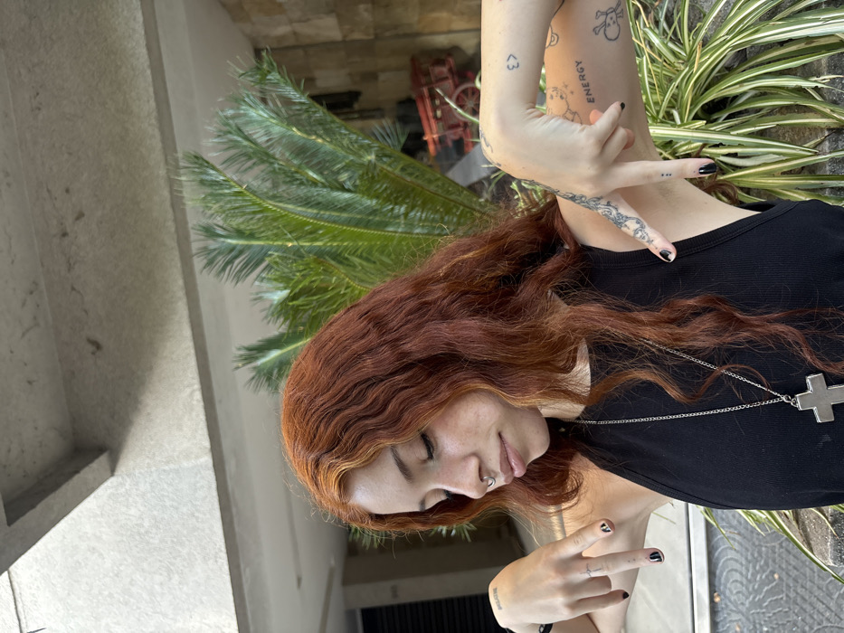 A woman with red, wavy hair is shown from the torso up, positioned among greenery. She is wearing a black tank top and a silver cross necklace. Her arms are raised, with visible tattoos on her forearms and fingers painted with black nail polish. A palm tree's leaves are behind her head, and a building is to her left. There are other plants with green and white striped leaves behind her right arm.