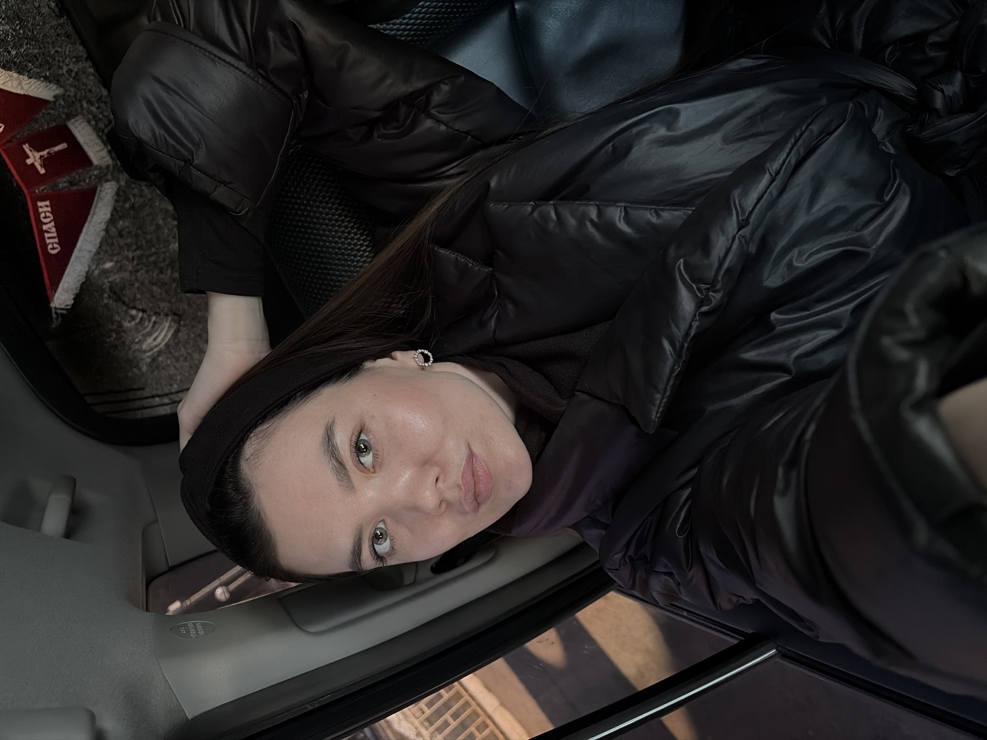 The image shows a woman wearing a black headband and a dark puffer jacket in the interior of a car. The view is angled, likely taken from a phone camera. A triangular pennant with text in Cyrillic and a cross is visible in the background. The setting is possibly in Chaikovsky, Russia.