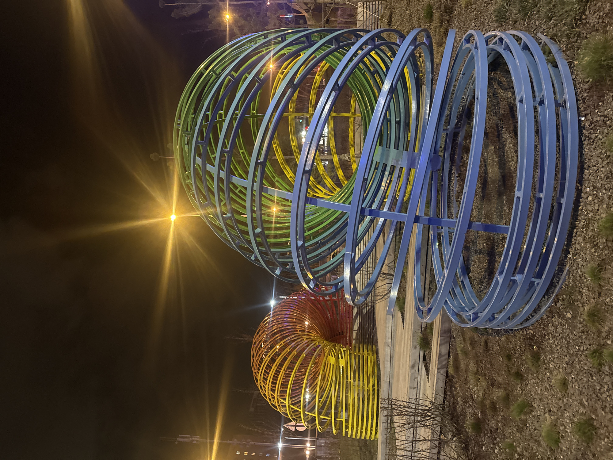 The image captures a nighttime scene in Yekaterinburg, Russia, featuring three colorful, metal tunnel-like sculptures. The sculpture in the foreground is predominantly blue, transitioning into green and yellow towards the center. A smaller sculpture composed of yellow and orange rings is visible in the background. The sculptures are situated on a gravelly ground surface, with sparse vegetation nearby. In the background, city lights illuminate the scene. The image appears to have been taken at night, under artificial lighting conditions.